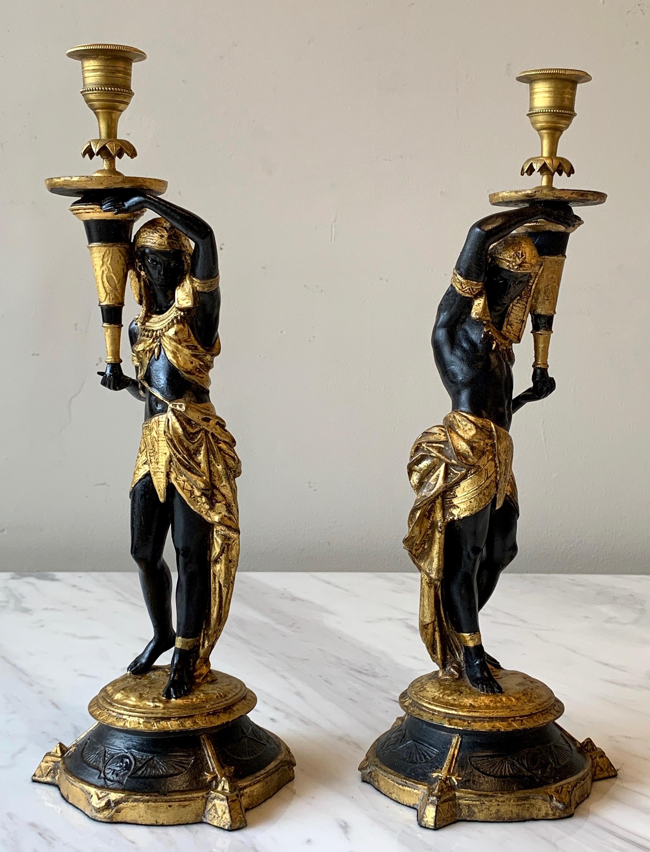 A fantastic mirrored pair of late 20th century. Egyptian revival ebonised and gilt decorated cast resin candlesticks depicting a male and female character holding torchieres.