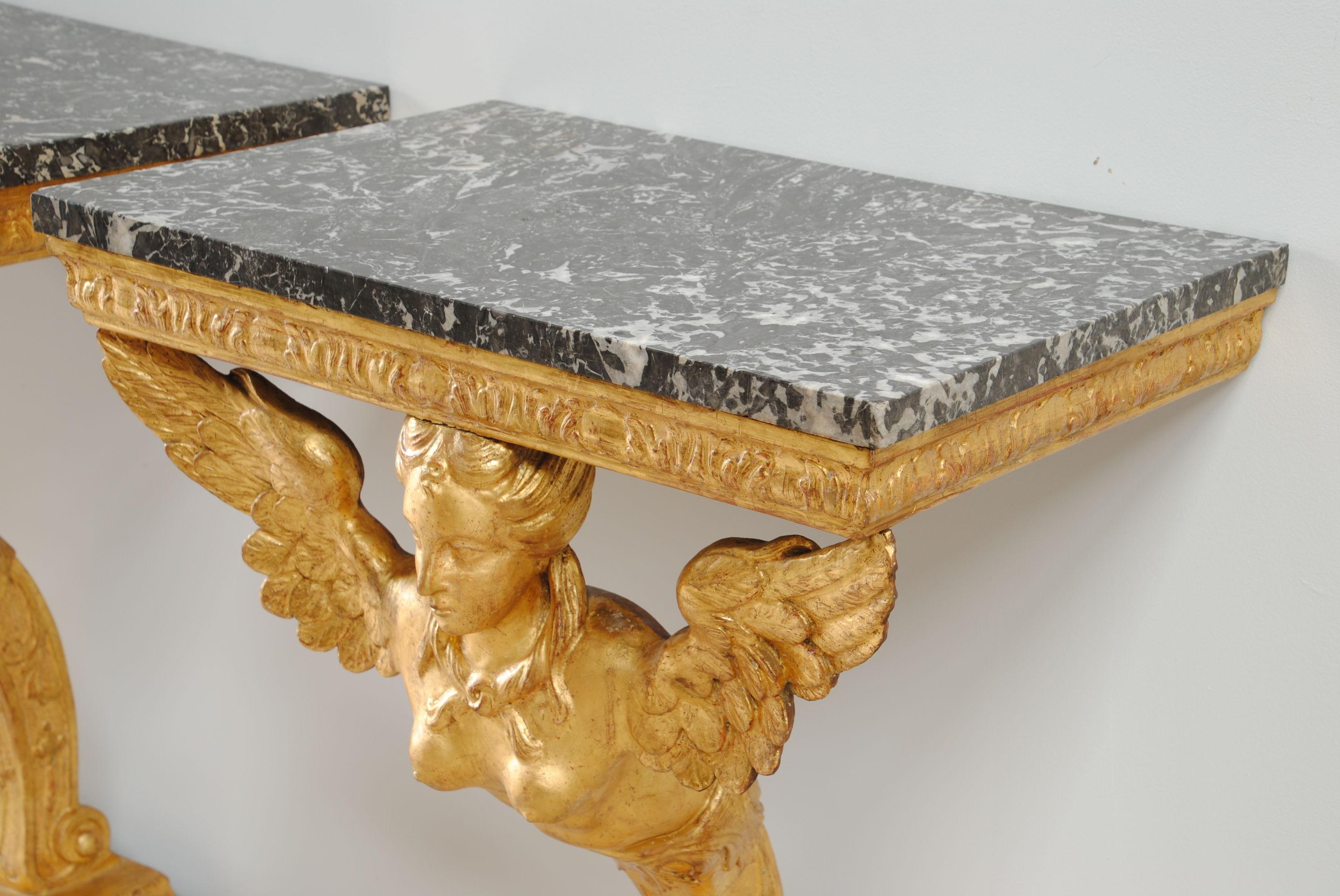 A pair of carved giltwood console tables with winged, busted maidens holding grey marble tops on a single kent design plinth.