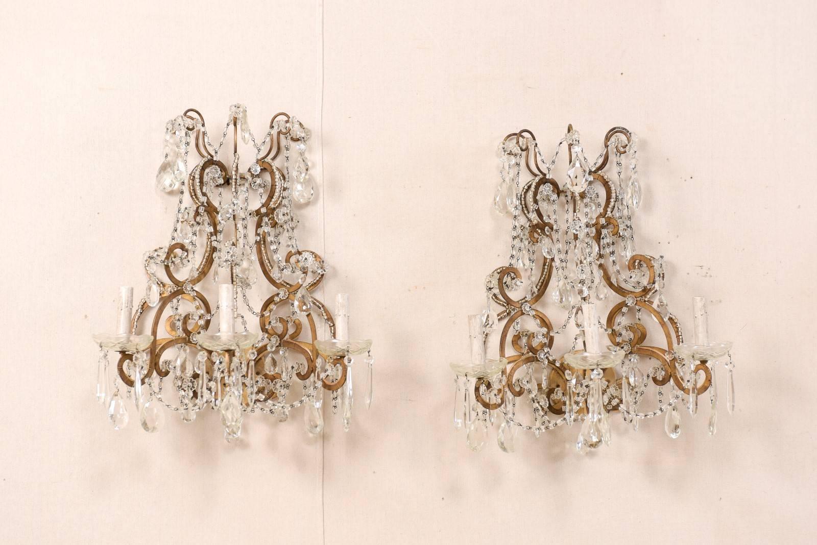 A pair of Italian mid-20th century crystal three-light wall sconces. This pair of Italian crystal sconces from the mid-20th century features a variety of faceted crystals ornately decorating the scrolled and gilded metal armature and arms. Strands