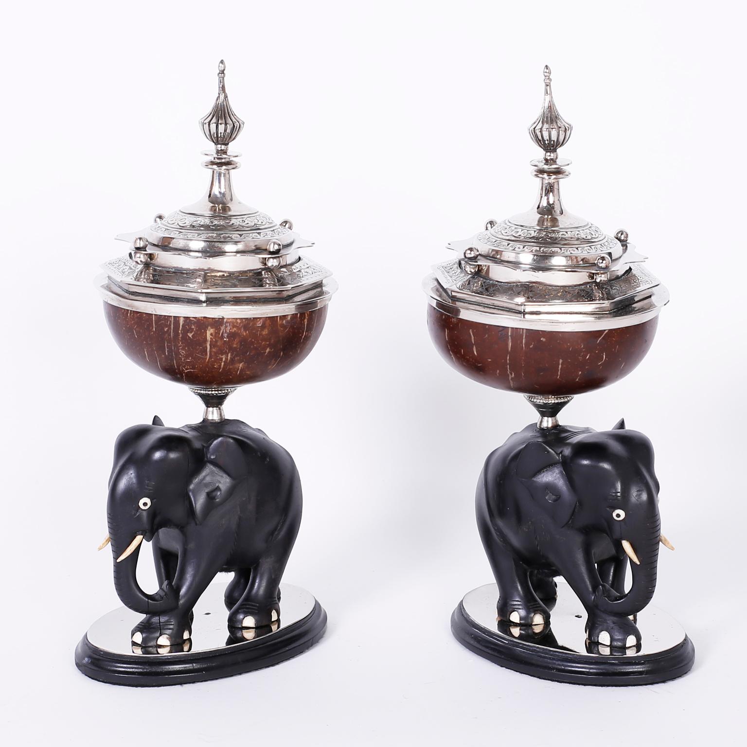Antique Anglo-Indian garniture boxes featuring silvered metal lids, dramatic finials and hand-hammered floral and geometric designs over a coconut vessel. The carved ebony elephants have bone eyes, tusks and toes, all this presented on metal and