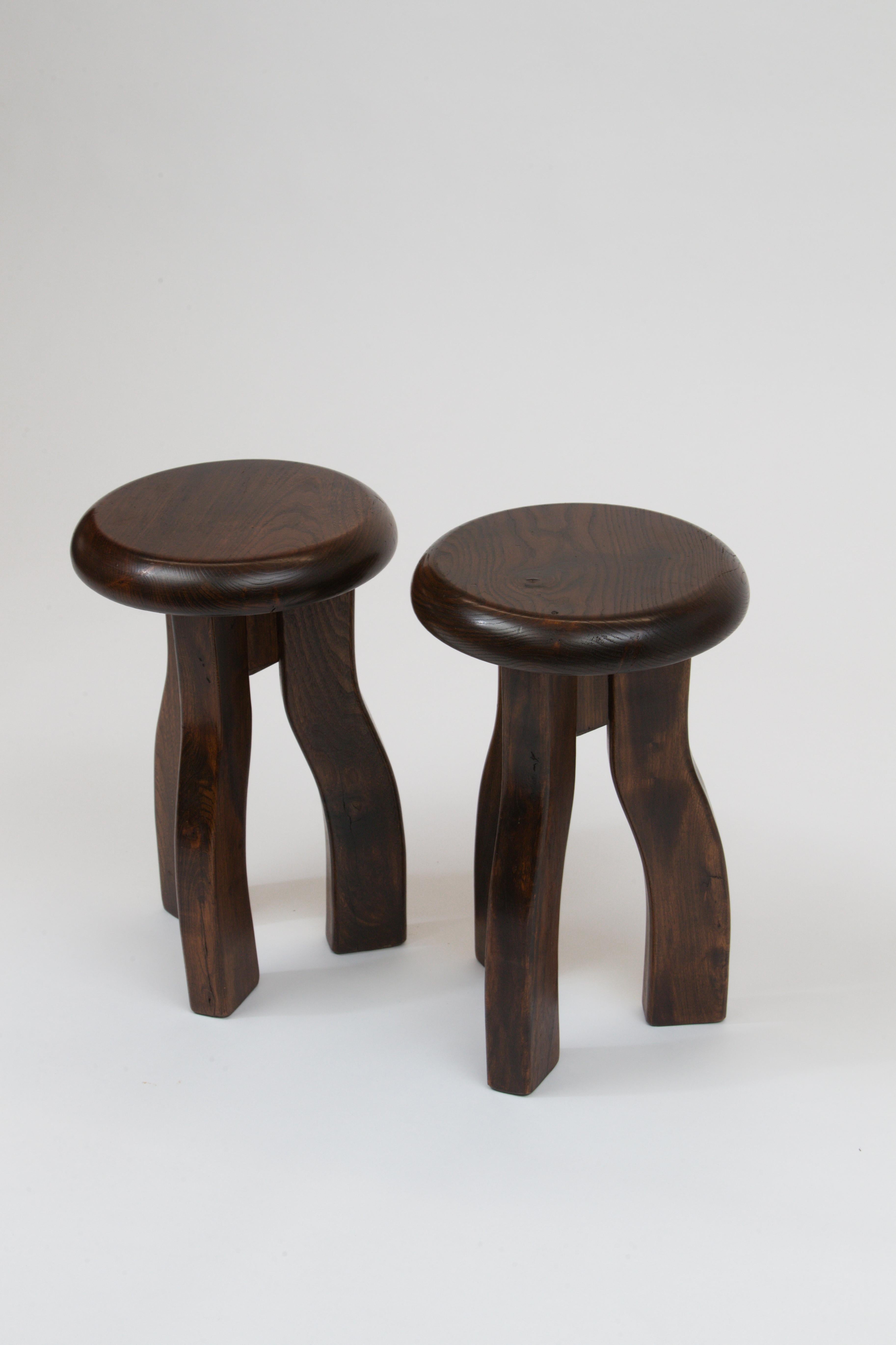 An unusual pair of three-legged stools with a solid top and thick curving legs, made of Elmwood and stained a rich brown.

Also nicely used as movable side tables.