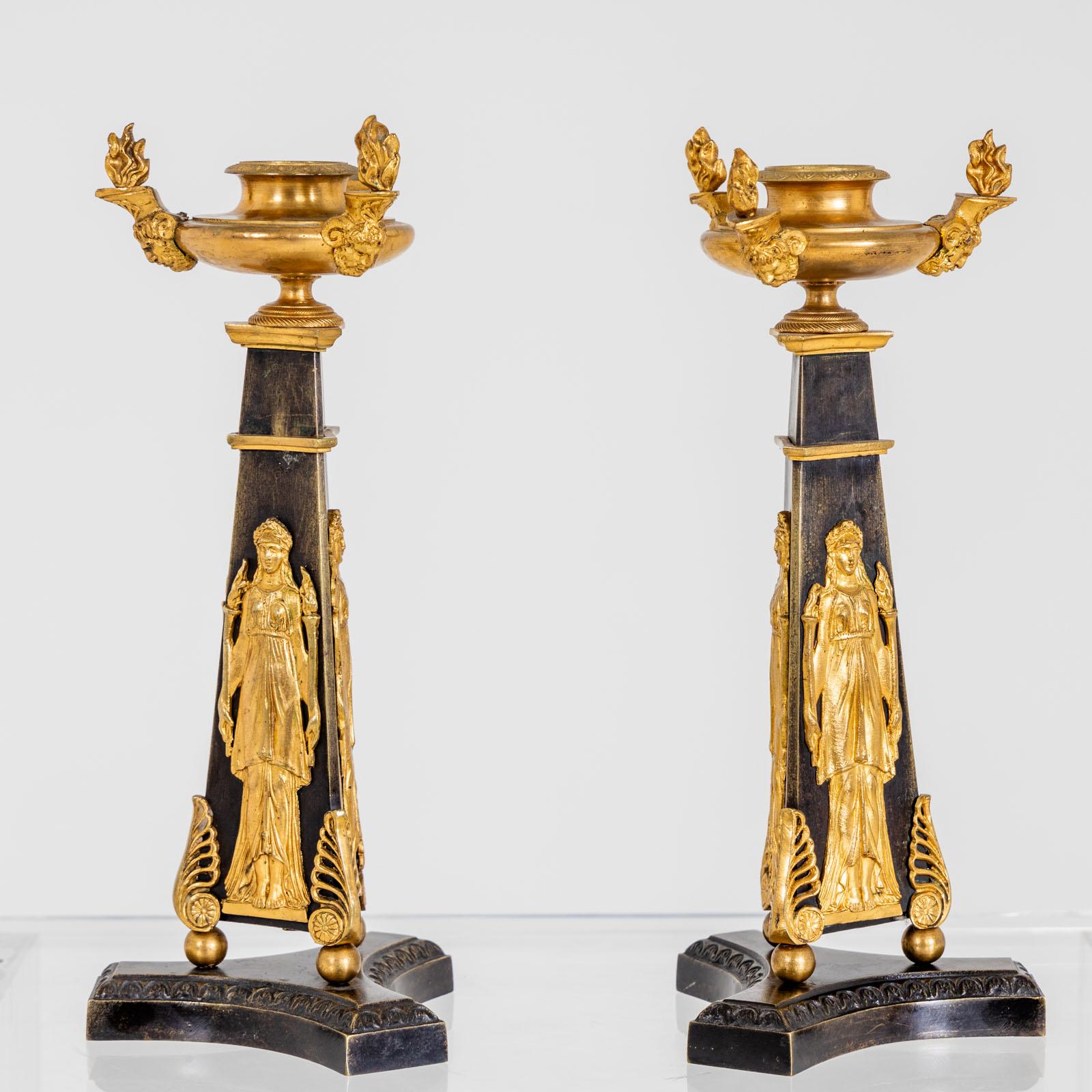 Coppia di candelieri in bronzo brunito e dorato a fuoco. I beccucci sono circondati da tre maschere di satiro coronate da fiamme. Il fusto piramidale è decorato con tedofori in rilievo. I candelieri si trovano sopra tre sfere su una base a