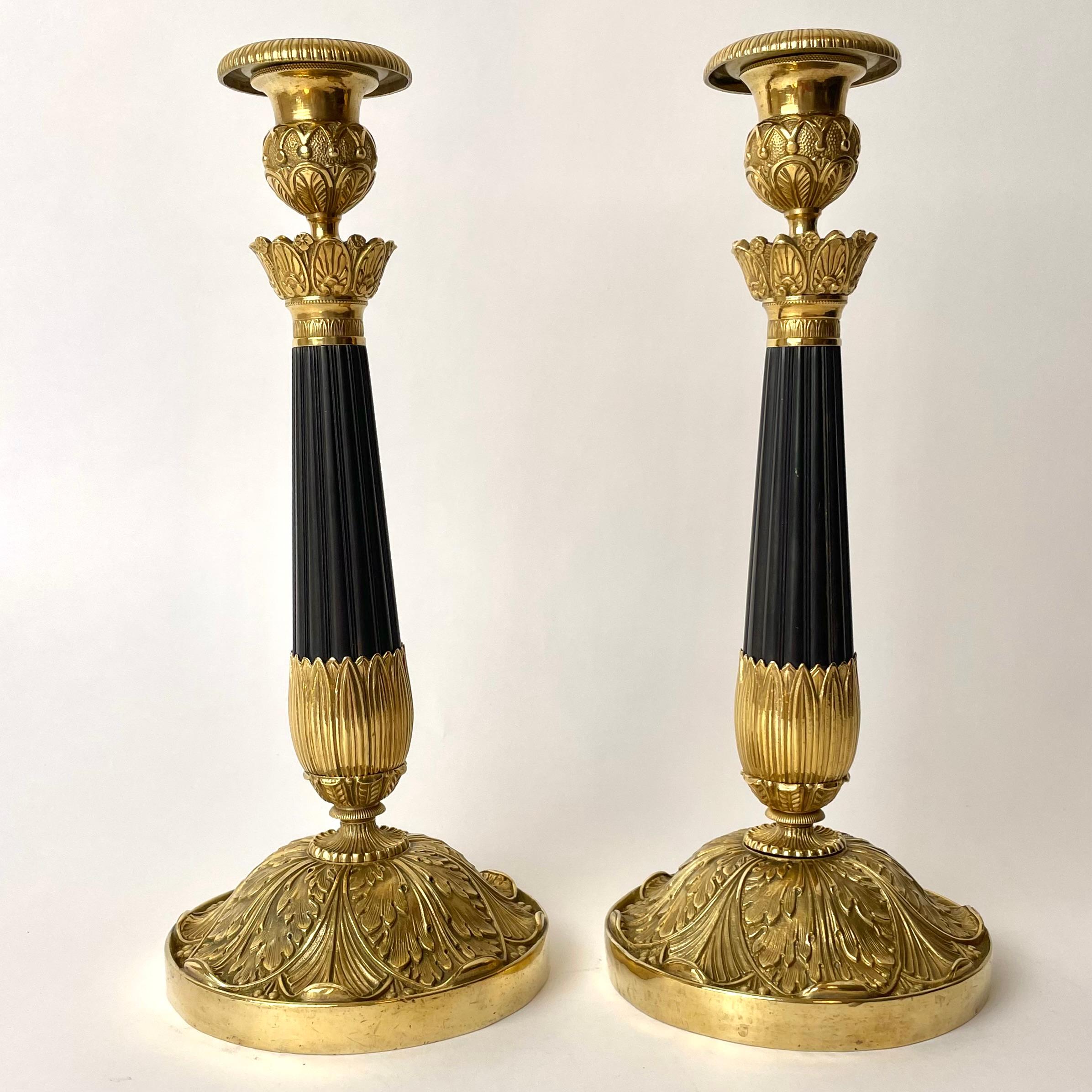 A Beautiful Pair of Empire Candlesticks, Gilded and Patinated Bronze. 

Richly ornamented base with flowing leaf-pattern. Dark patinated fluted column which then continues into a gilded bulb, which serves as a base for the cup. Made in France,
