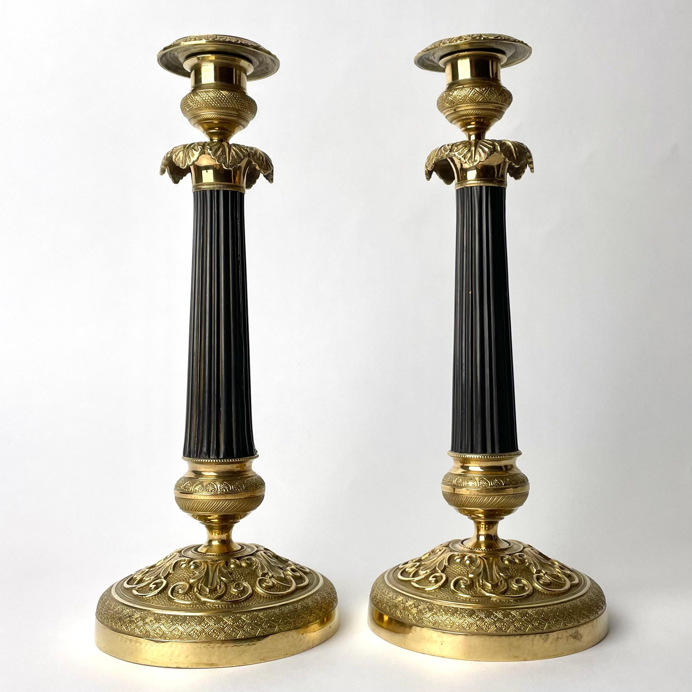  A beautiful Pair of Empire Candlesticks in gilded and darkpatinated bronze. Made in France during the 1820s. Richly decorated with leaves and other period ornaments as well as a dark patinated column in the center of the candlestick.

Wear