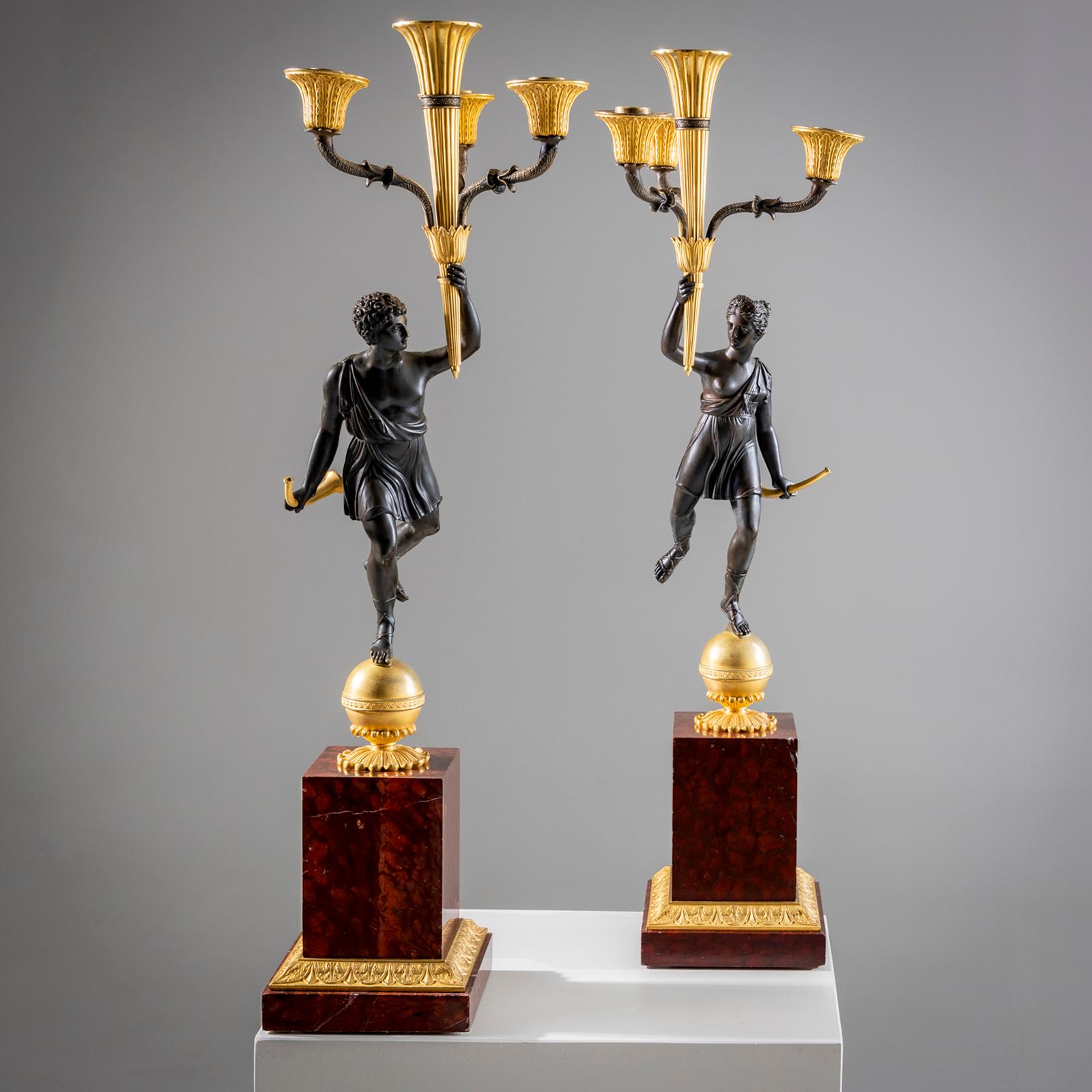 Coppia di girandole francesi in bronzo brunito e in parte dorato a fuoco, su una base in marmo rosso con decorazione sferica. Le figure dei candelabri, disegnate come Apollo e Diana, portano i candelabri a quattro fiamme in una mano e i corni da