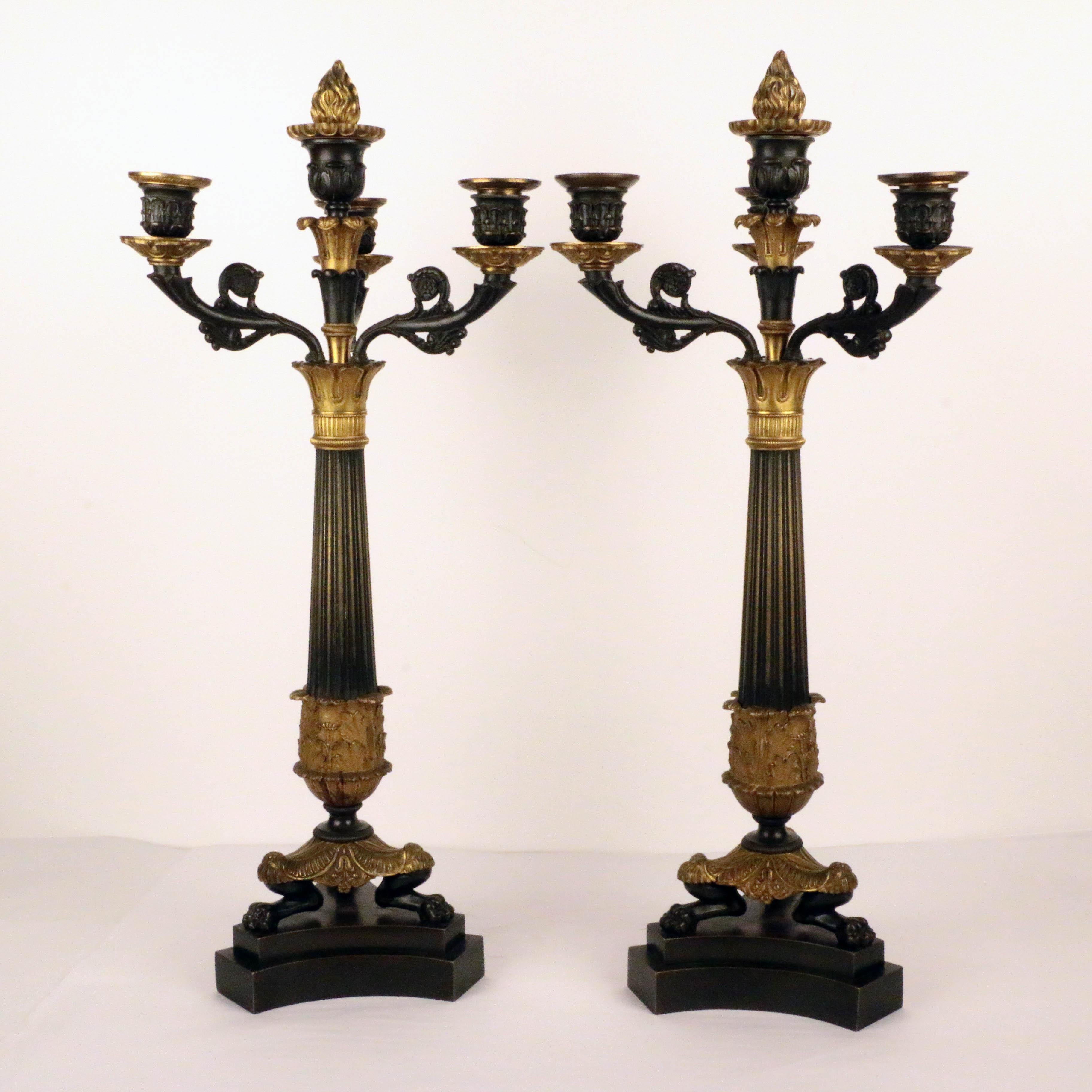Coppia di candelabri a quattro luci di epoca Impero, su zampe di leone a treppiede su un basamento incurvato a gradini, che sostengono una base a palmetta con colonna ribattuta e stelo centrale della candela dorato; lo spegnimento a fiamma