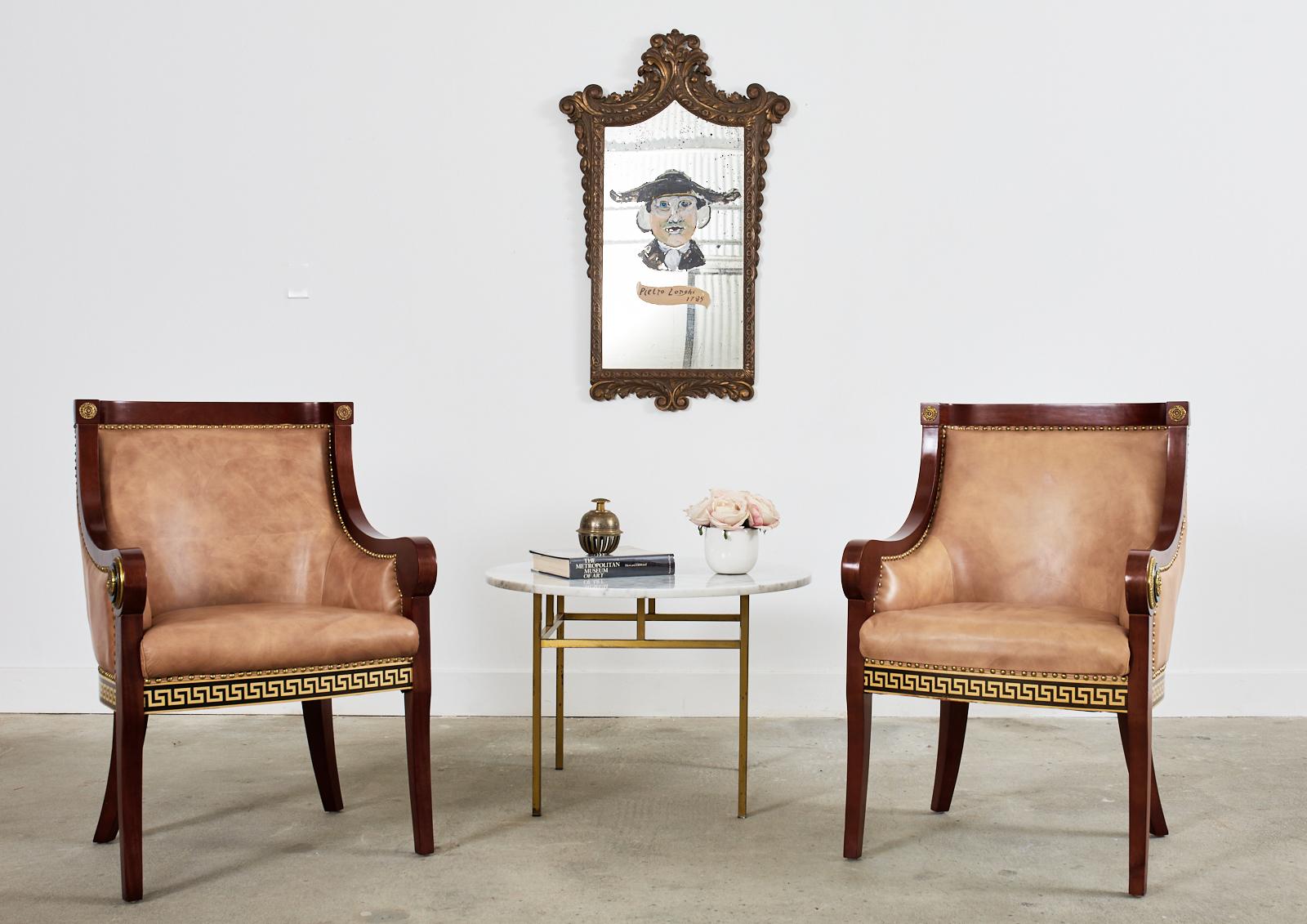 Pair of maximalist empire revival style armchairs featuring Versace inspired motifs. The chairs have a classic gondola shaped frame with faux leather naugahyde upholstery. The faux leather is bordered by shiny brass tack nail heads. The chairs are