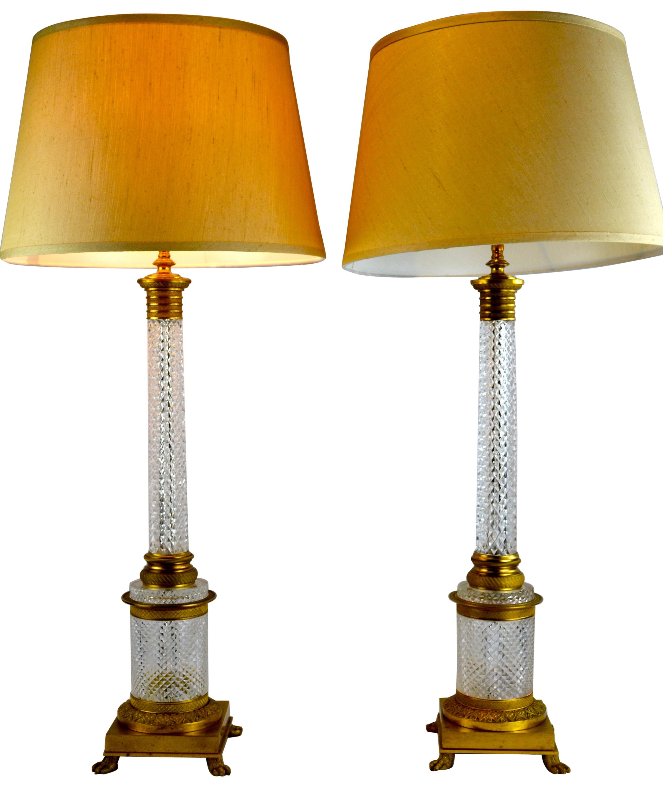 A pair of tall lamps featuring cut crystal columns with gilt bronze capitals and bases that in turn are supported on wider cut crystal and gilt bronze drum shaped bases terminating in a square gilt bronze bases with lions paw feet.