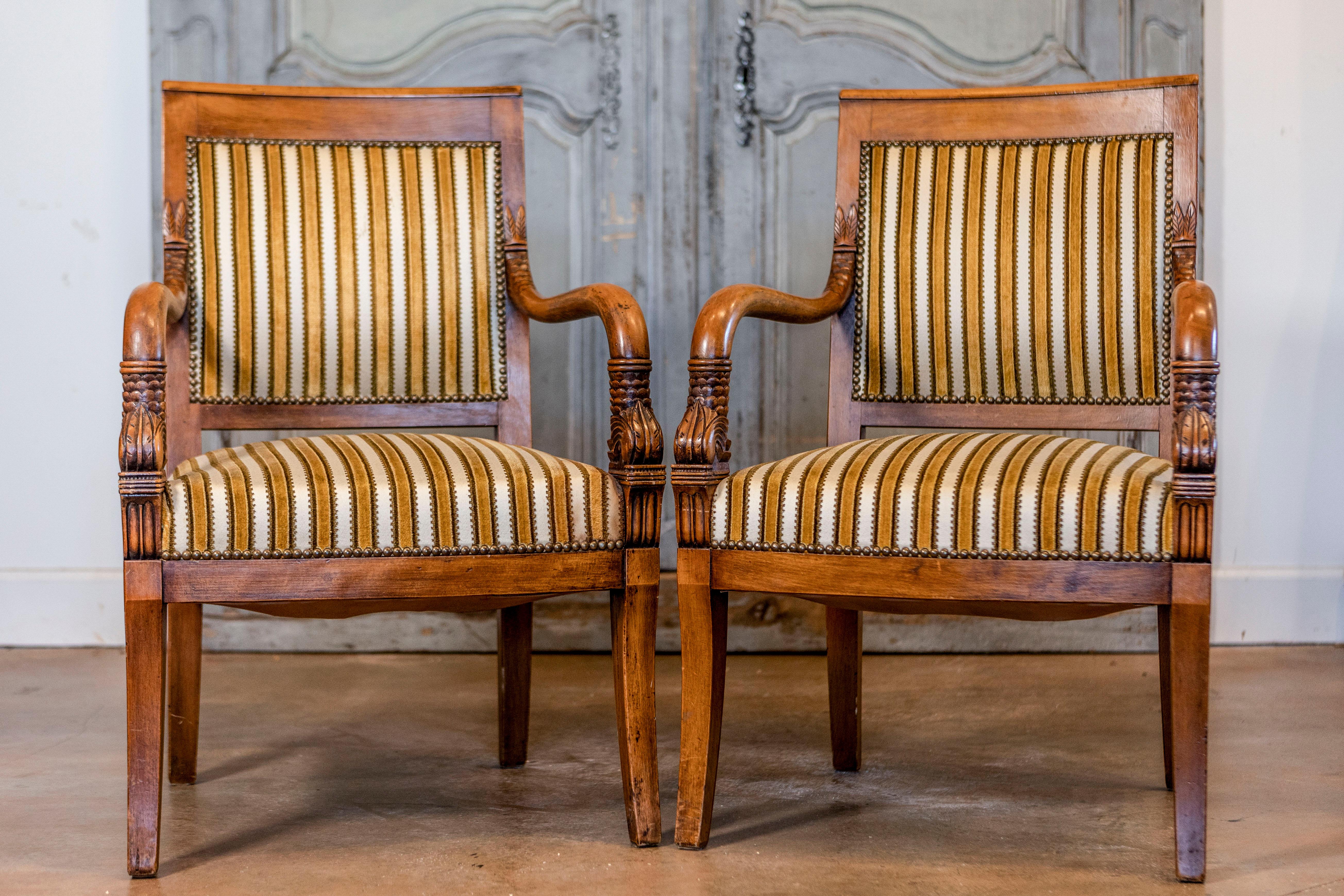 A pair of French Empire style light walnut armchairs with foliage carved curving arms and saber legs. Step back in time with these exquisite French Empire style armchairs, beautifully crafted in the 20th century. Featuring the iconic foliage-carved