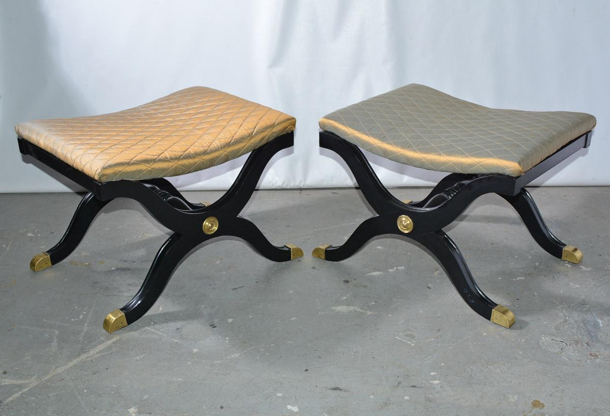 The pair of 20th century Empire-style Curule benches are painted black with protective brass feet and escutcheons. The center bar is turned wood. The padded seats are newly upholstered in iridescent gold quilted silk fabric. 

Measures: Middle