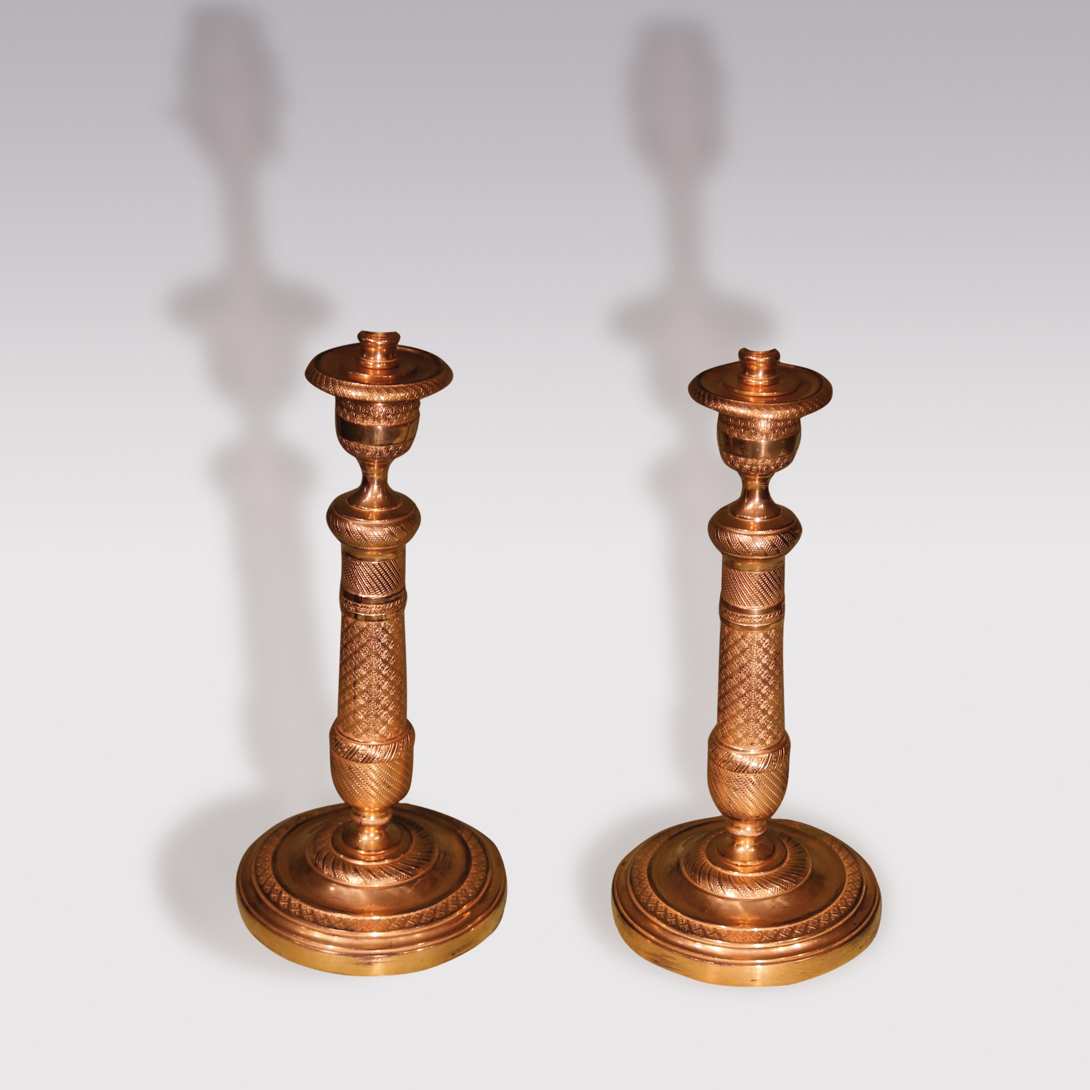 A pair of early 19th century ormolu candlesticks, engine -turned throughout, having urn-shaped nozzles above decorated tapering stems supported on circular bases. (Now converted to lamps).