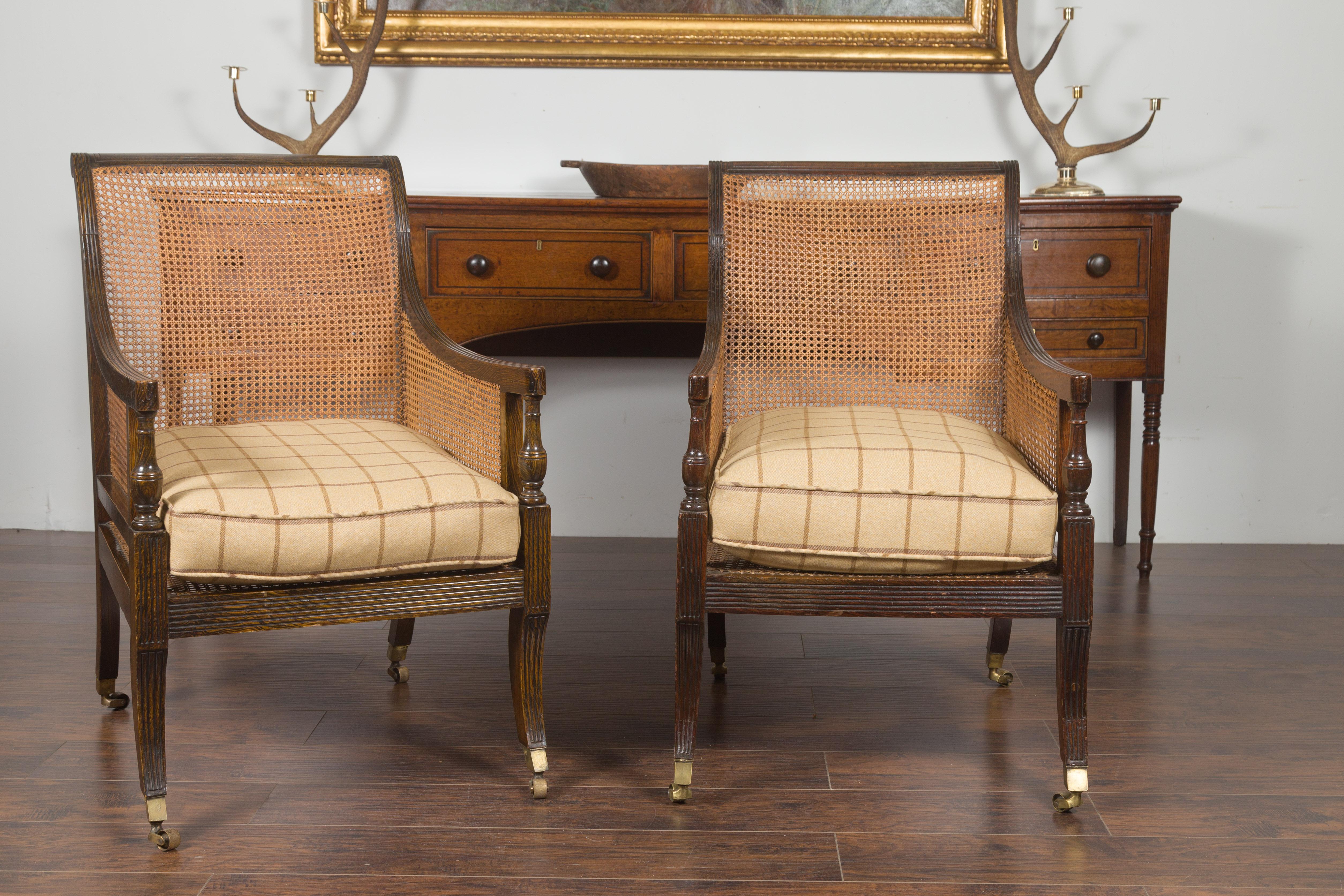 A pair of English oak library chairs from the late 19th century, with cane and custom cushions. Created in England during the third quarter of the 19th century, this pair of cane and oak library chairs features out-scrolling backs flowing seamlessly