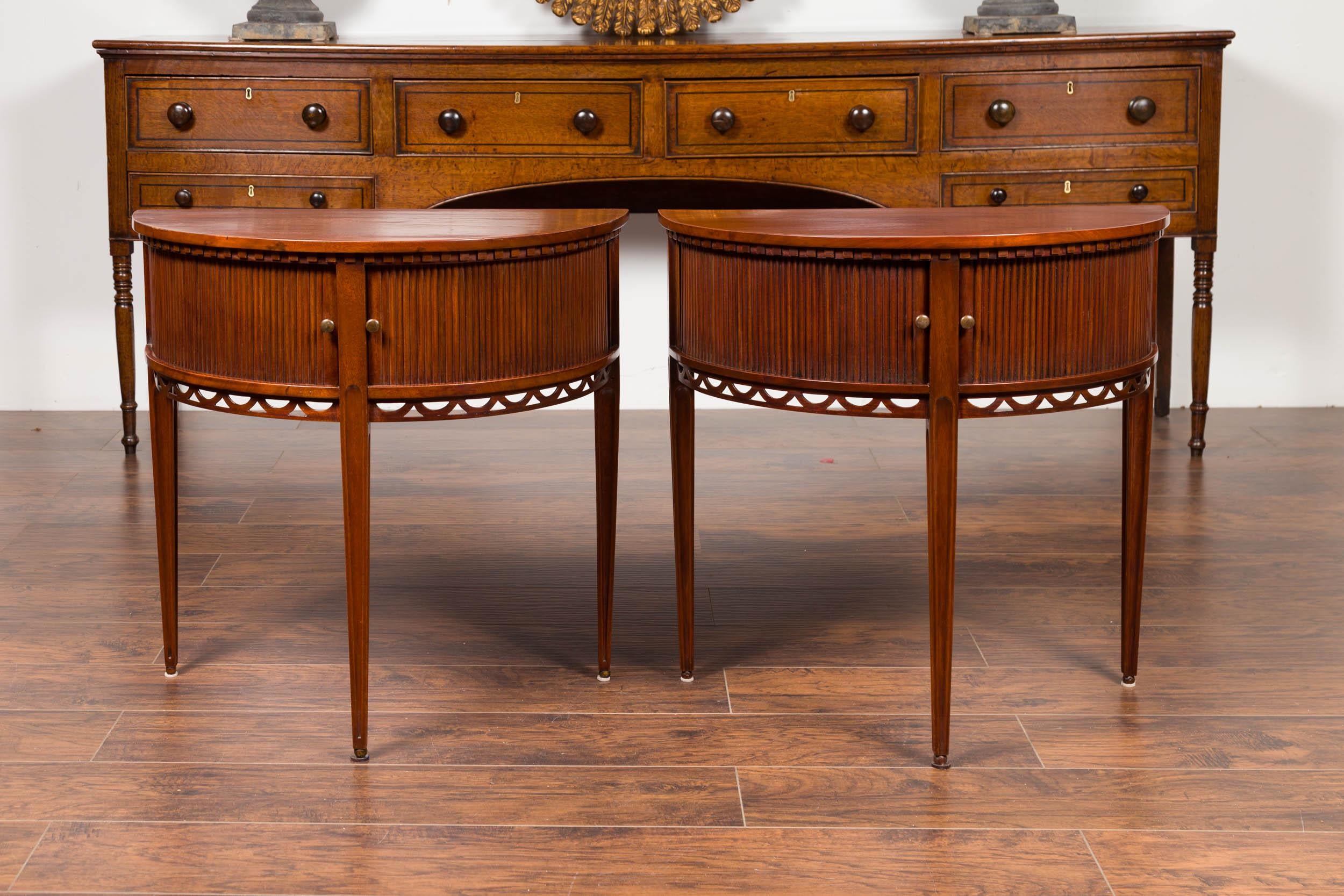 A pair of English mahogany demilune tables from the late 19th century, with reeded sliding doors and carved apron. Born in England during the third quarter of the 19th century, each of this pair of demilune features a semi-circular top sitting above
