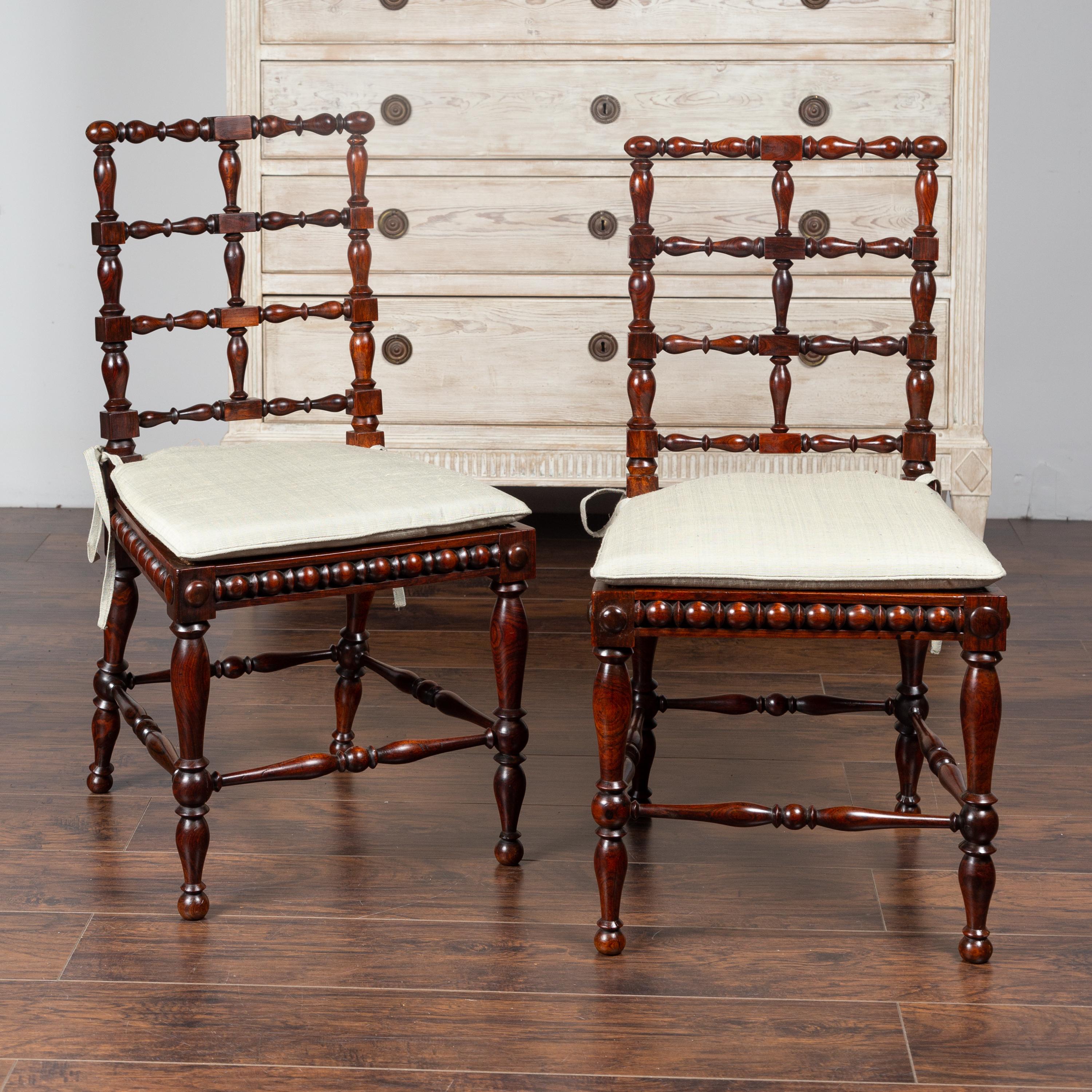 A pair of English rosewood side chairs from the 19th century, with turned spindle motifs, cane seats and small cushions. Born in England during the third quarter of the 19th century, each of this pair of side chairs features a slightly slanted back