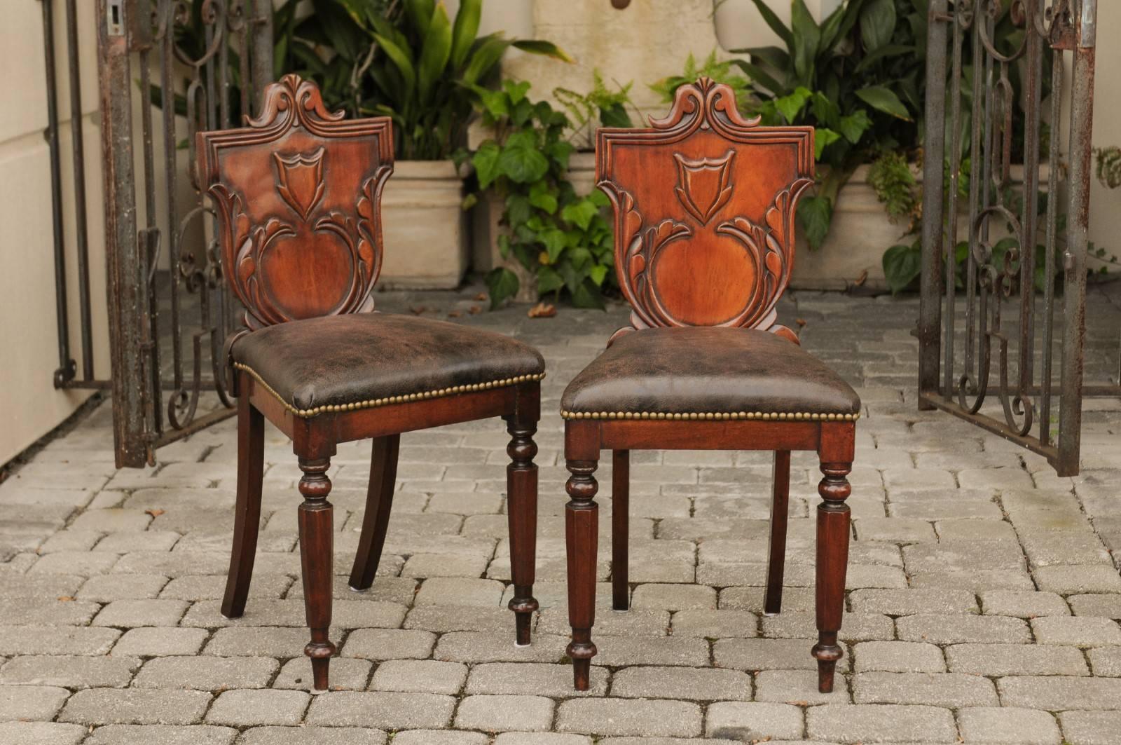 A pair of English carved oak hall chairs from the late 19th century with carved shield-shaped backs and leather upholstered seats. Each of this pair of English hall chairs features an exquisite back, carved in the shape of a shield and adorned with