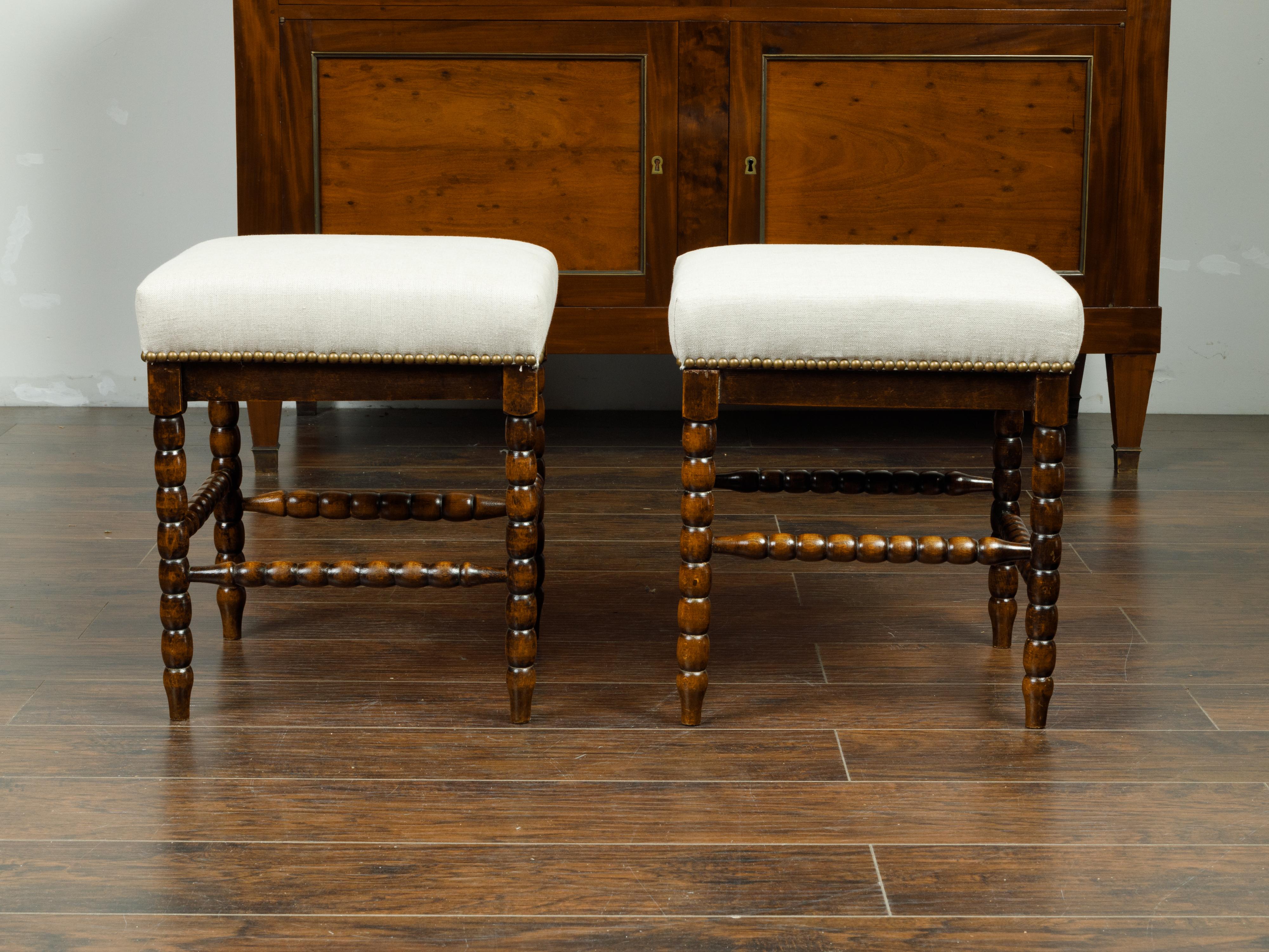 A pair of English wooden bobbin stools from the late 19th century, with side stretchers and new upholstery. Created in England during the last quarter of the 19th century, each of this pair of stools features a square top newly reupholstered with a