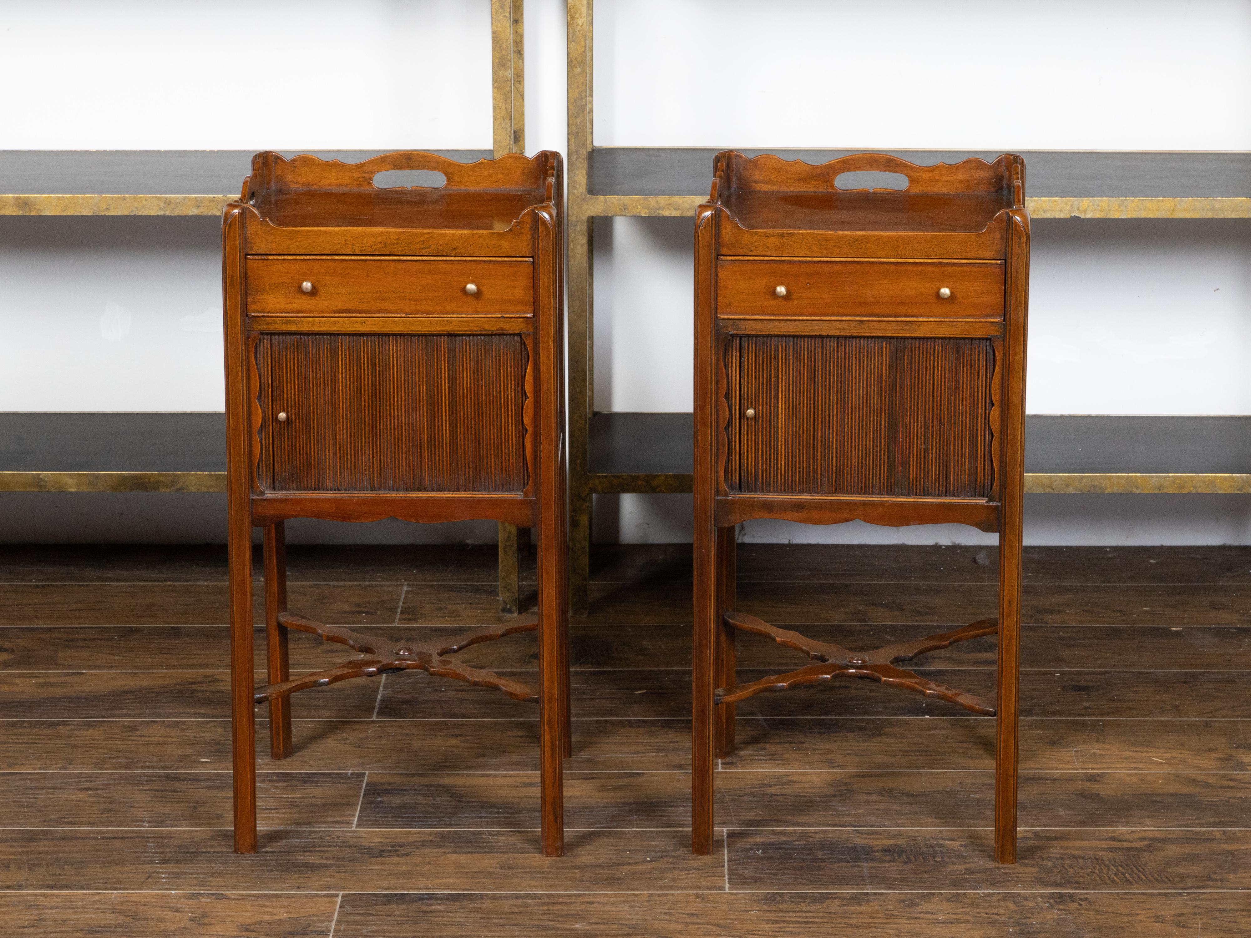A pair of English mahogany bedside tables from the early 20th century, with tray top, single drawer, tambour door and carved X-Form stretcher. Created in England during the turn the century that saw the transition of the 19th to the 20th century,