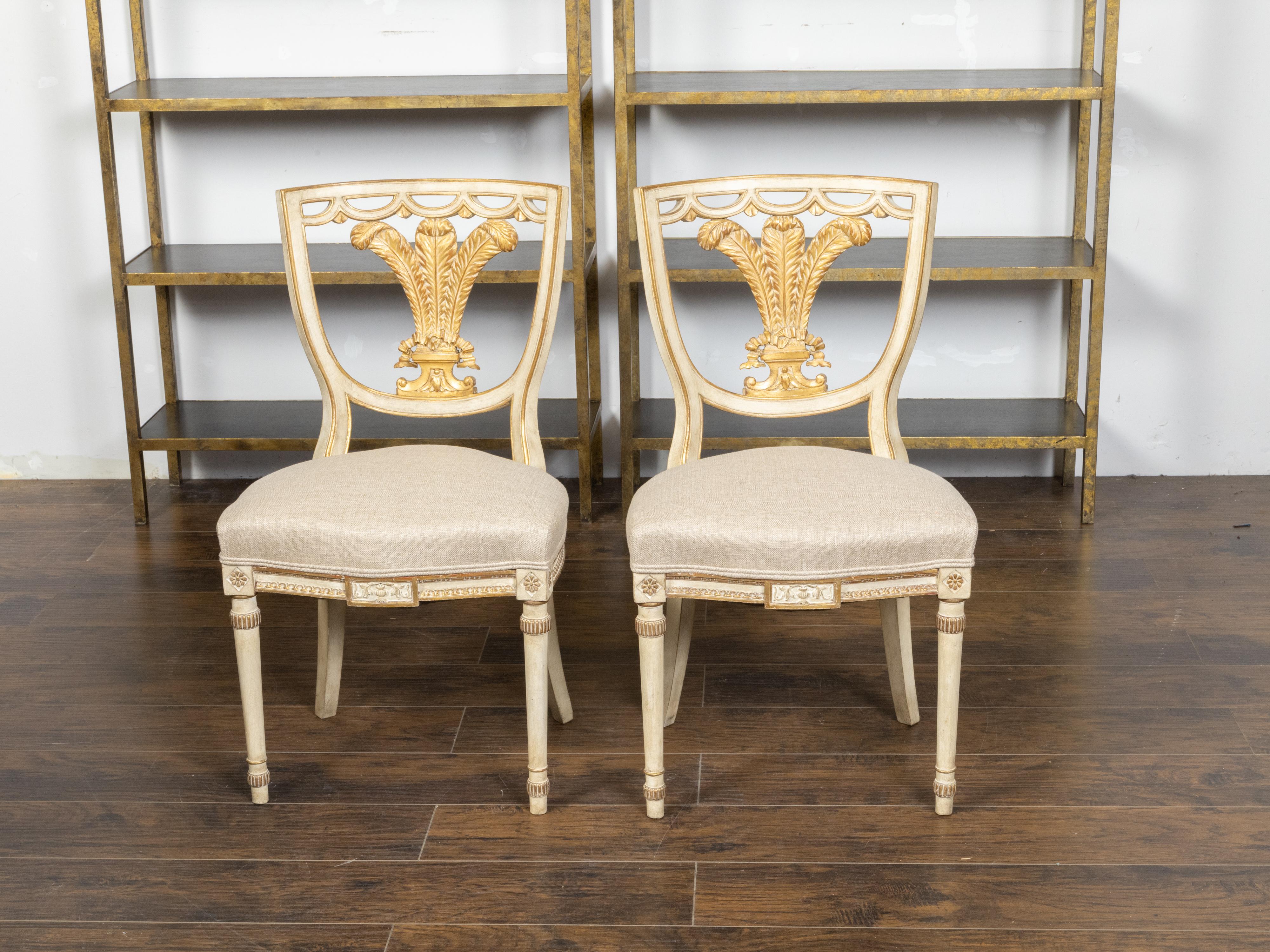 A pair of English Neoclassical style painted wood side chairs from the early 20th century with parcel gilt décor and carved feather motifs. Created in England at the Turn of the Century which saw the transition between the 19th to the 20th, this