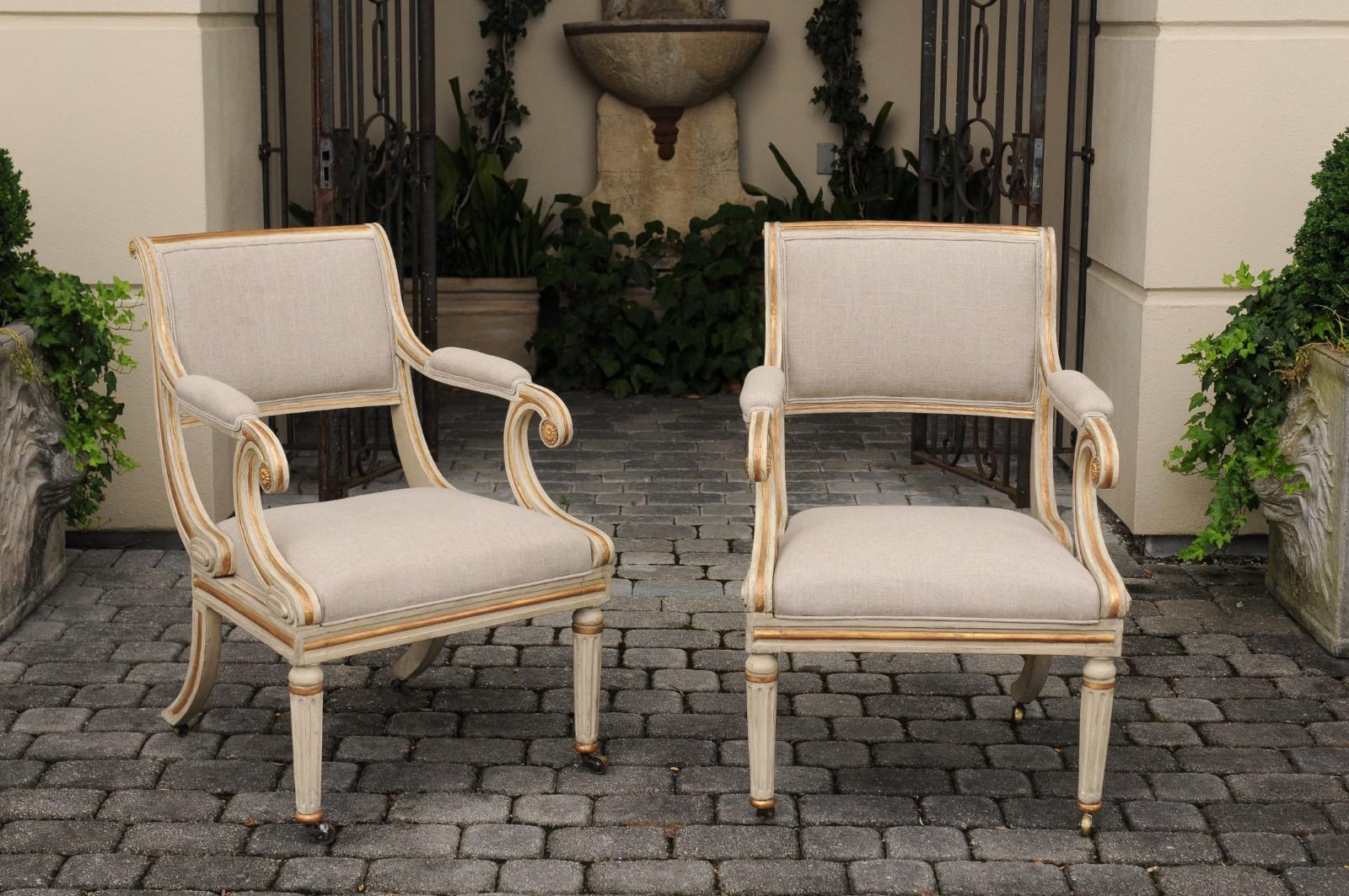 A pair of Regency style English painted wood parcel-gilt armchairs from the early 20th century, with scrolling arms, rosettes, fluted and saber legs, casters and new upholstery. Born in England at the Turn of the century, each of these two armchairs