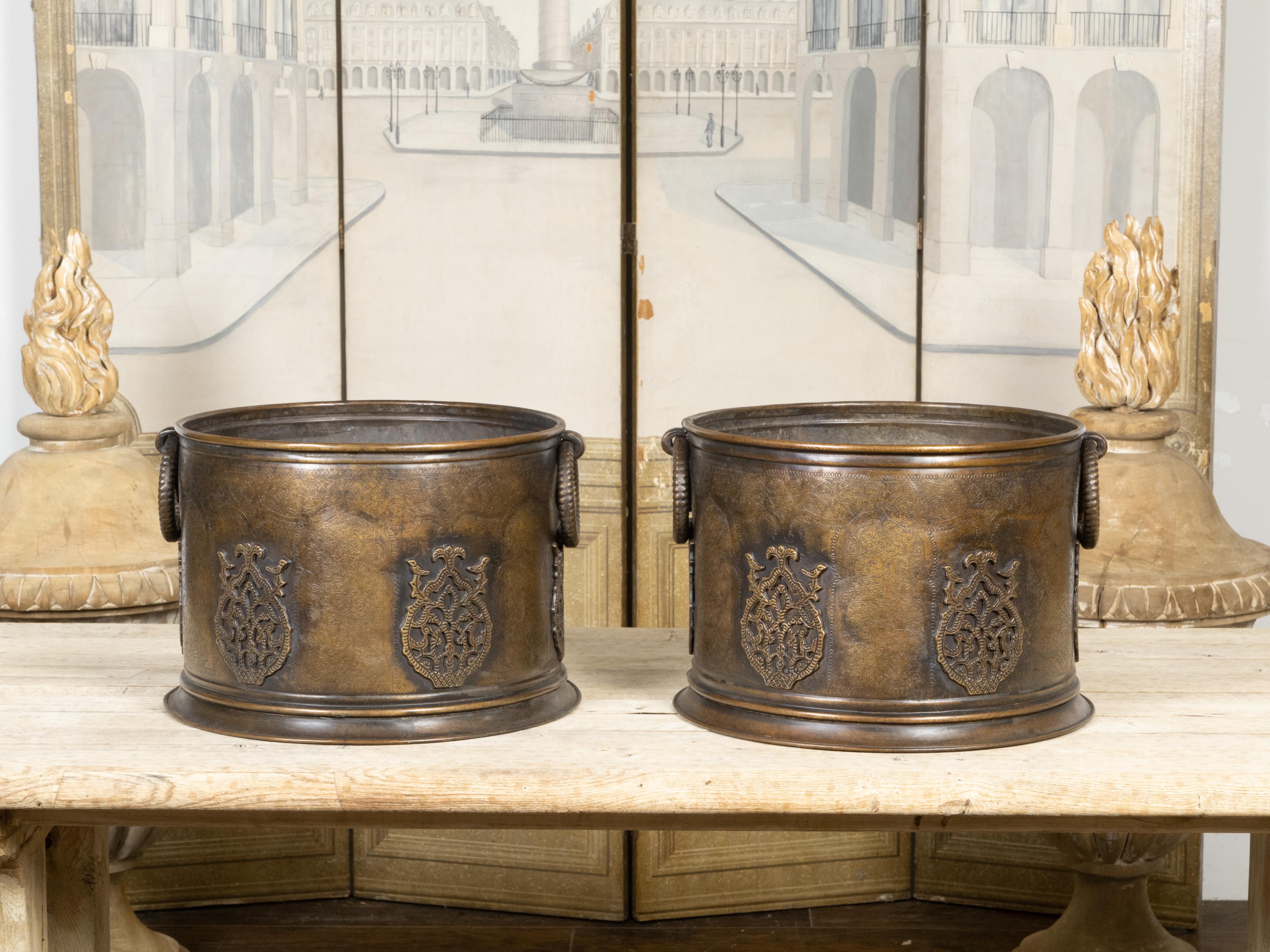 Une paire de cache-pots en cuivre anglais du début du 20e siècle, avec des poignées à anneau, un feuillage gravé et des motifs en relief. Créée en Angleterre au cours du premier quart du XXe siècle, cette paire de cache-pots en cuivre présente une