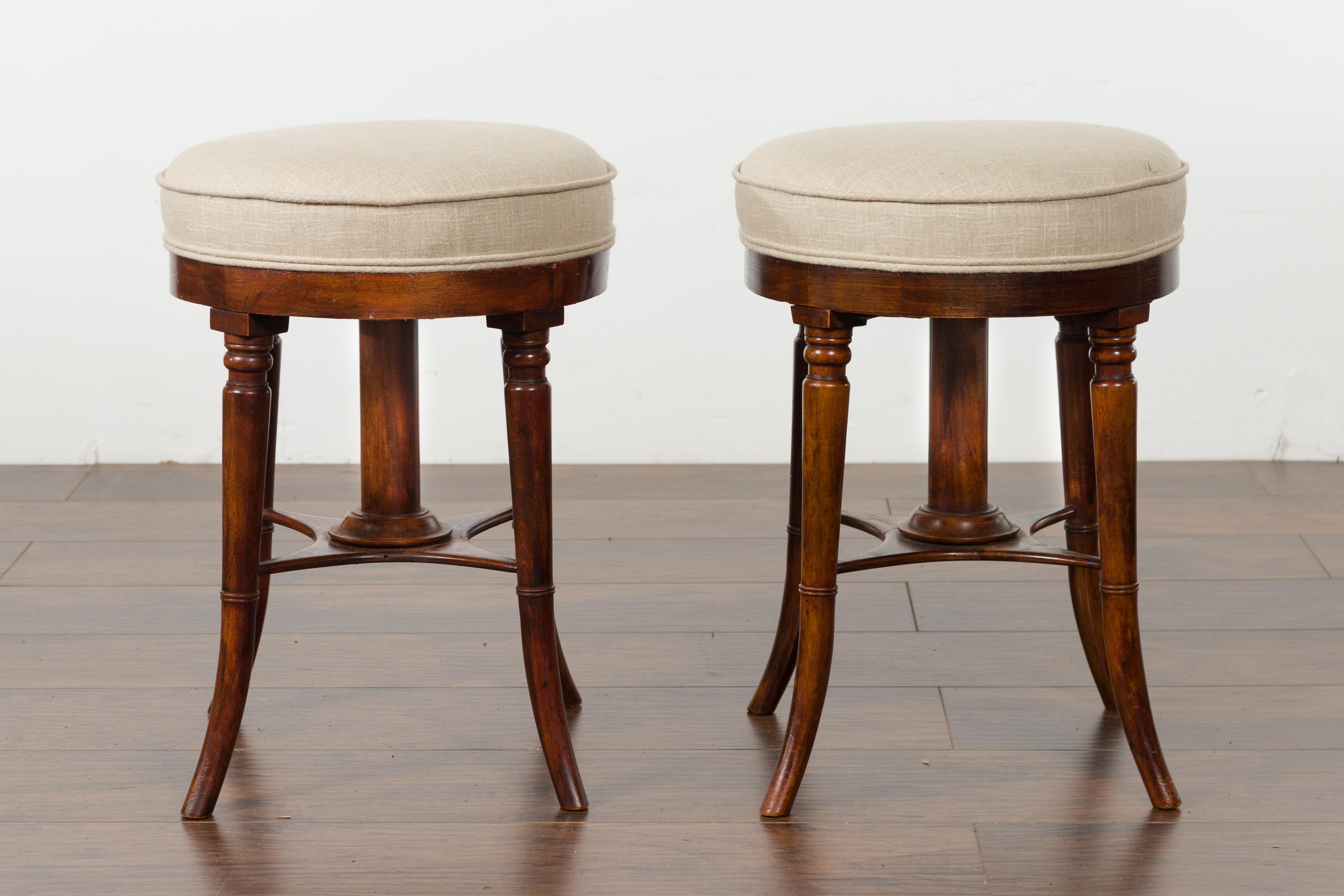 A pair of English mahogany stools from the early 20th century, with turned saber legs and new upholstery. Created in England during the first quarter of the 20th century, each of this pair of mahogany stools features a circular top, newly
