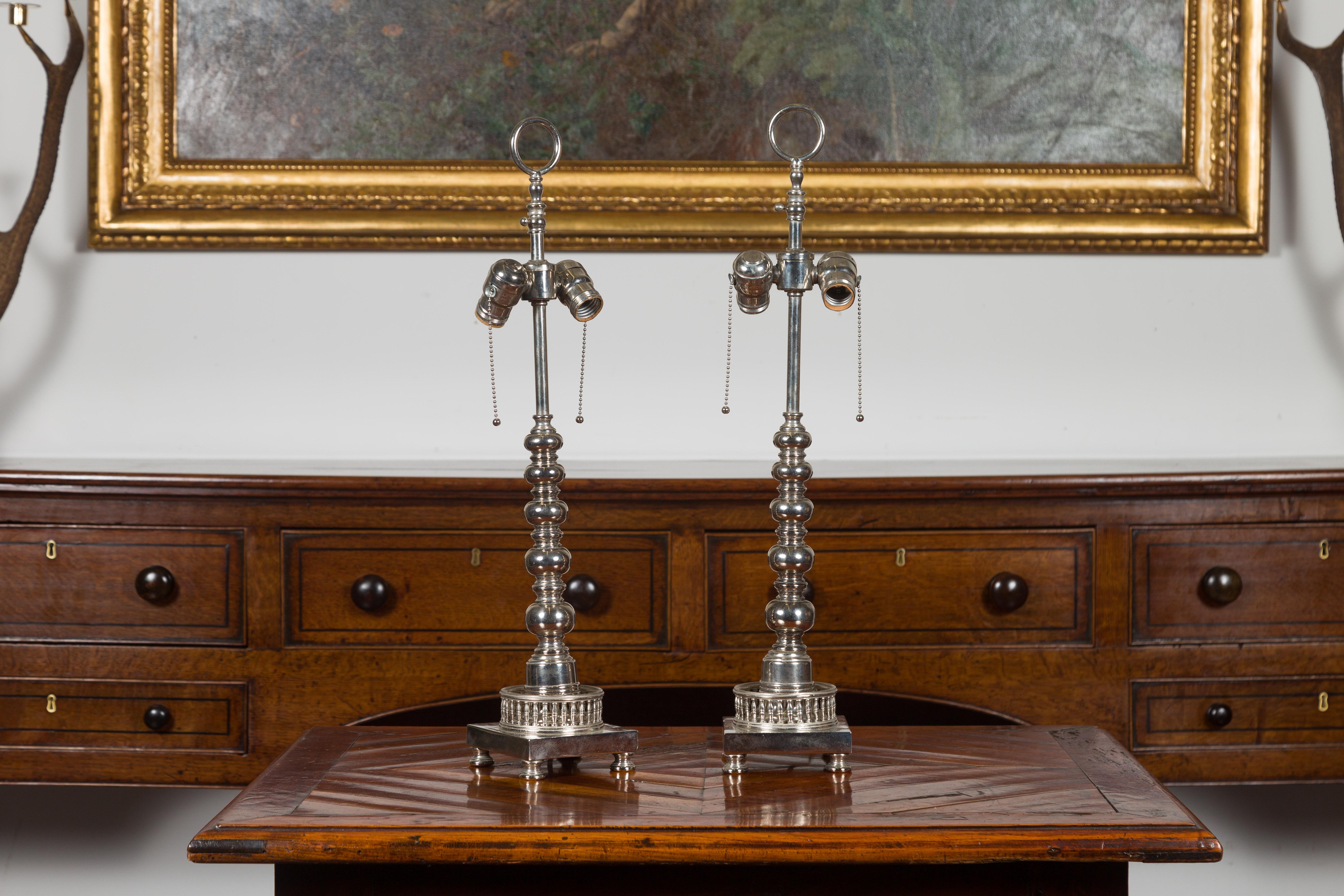 Une paire de lampes de table à deux lumières en métal argenté de style anglais, datant du milieu du 20e siècle, avec des accents de bobine et une galerie percée. Créée en Angleterre au cours du deuxième quart du XXe siècle, cette paire de lampes de