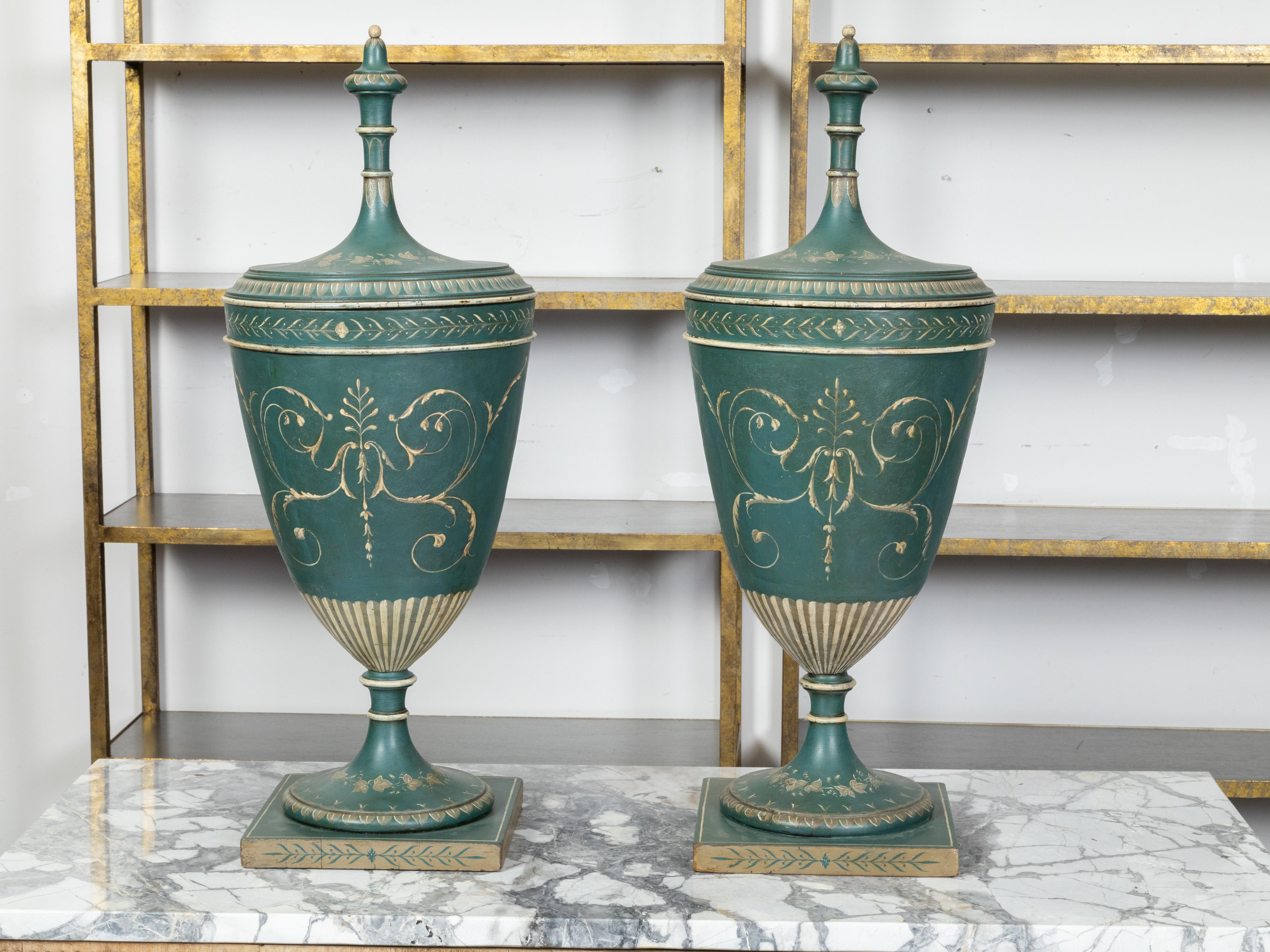 A pair of English Neoclassical green painted tôle lidded urns from the 19th century, with scrolling foliage and palmettes, friezes of waterleaves, campanula and ivy. Created in England during the 19th century, each of this pair of urns captures our