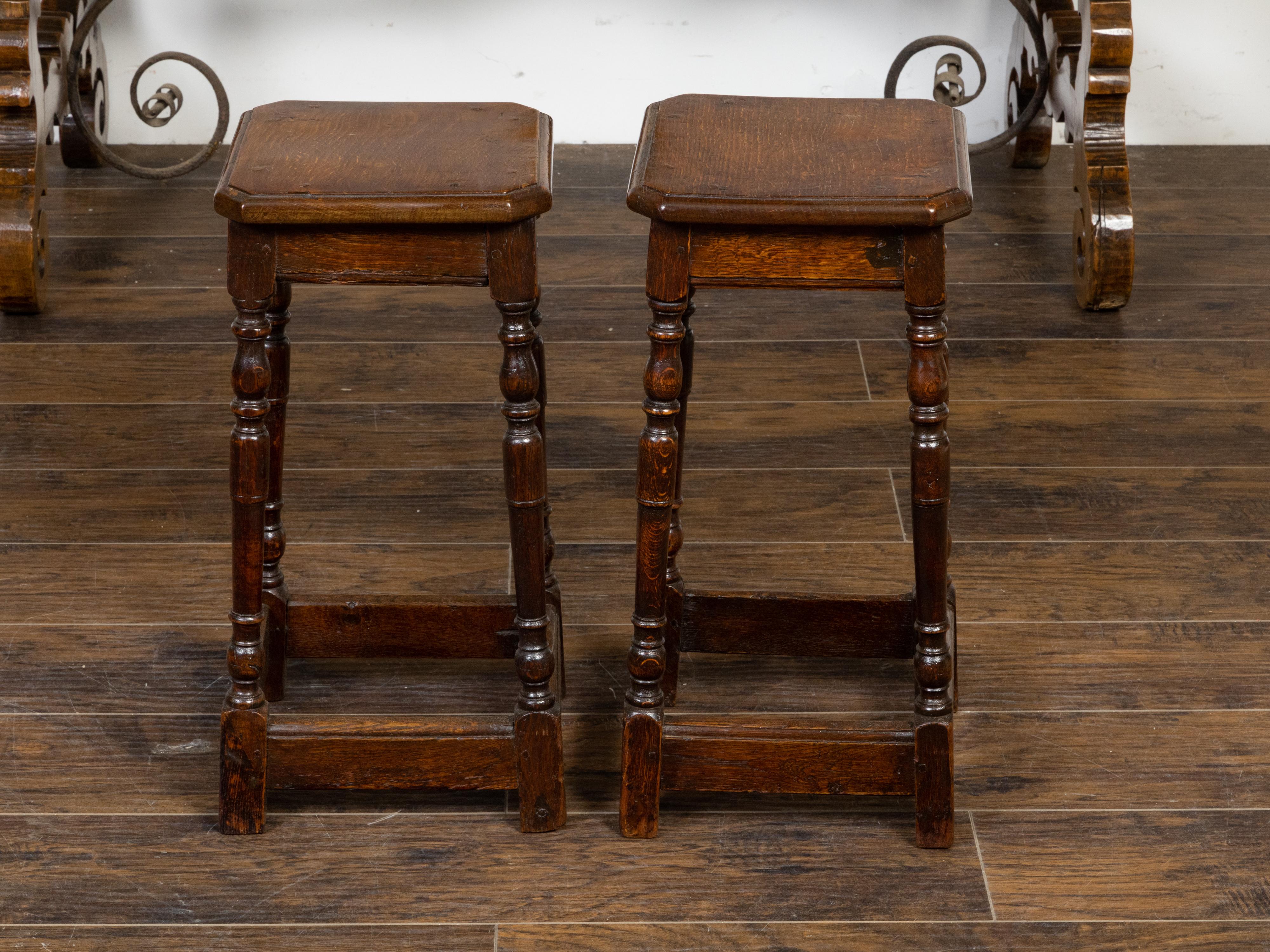 A pair of English oak joint stools from the 19th century, with square tops, turned legs and side stretchers. Created in England during the 19th century, each of this pair of joint stools features a square top with canted corners, sitting above a