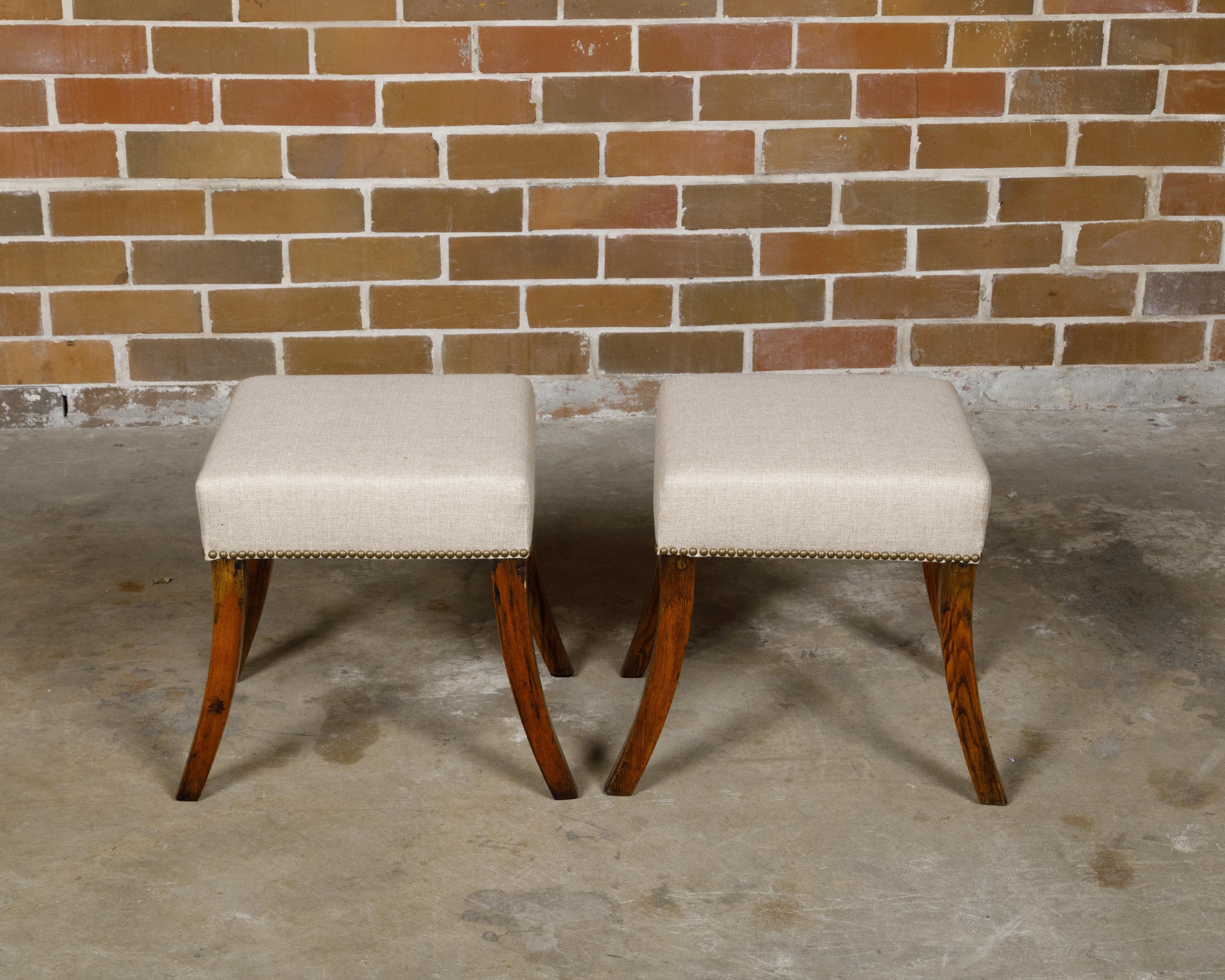 A pair of English oak stools from the 19th century with saber legs and custom upholstery. This pair of English oak stools from the 19th century is a splendid blend of traditional design and modern craftsmanship. Each stool stands on elegantly curved