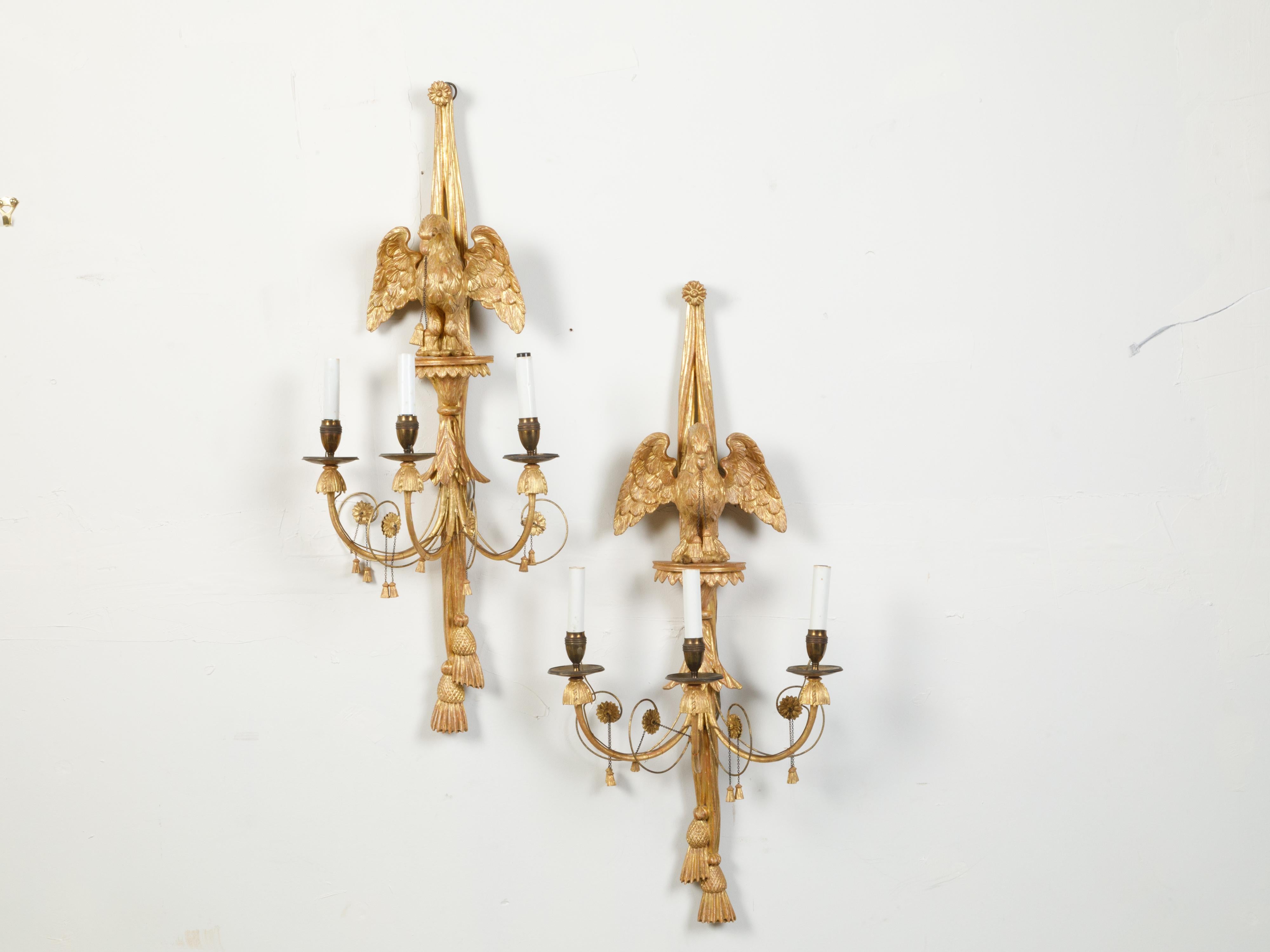 A pair of English giltwood sconces from the 19th century, with three lights and carved eagle motifs. Created in England during the 19th century, each of this pair of giltwood sconces captures our attention with its ribbon-tied eagle standing on a