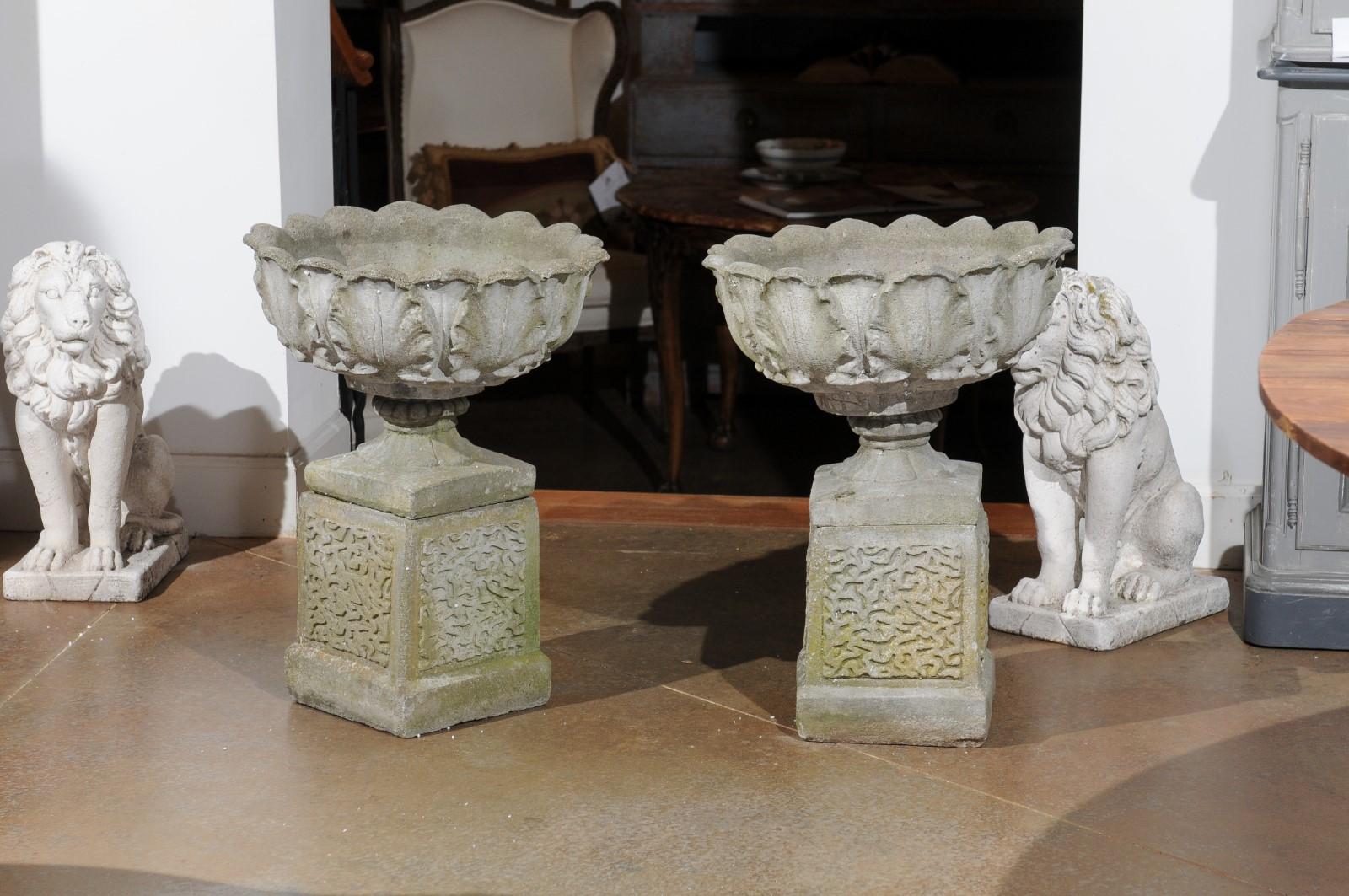 A pair of English carved stone urns on pedestals from the 20th century, with acanthus leaf motifs. Created in England during the 20th century, each of this pair of stone urns captures our attention with its elegant rhythm of acanthus leaves adorning
