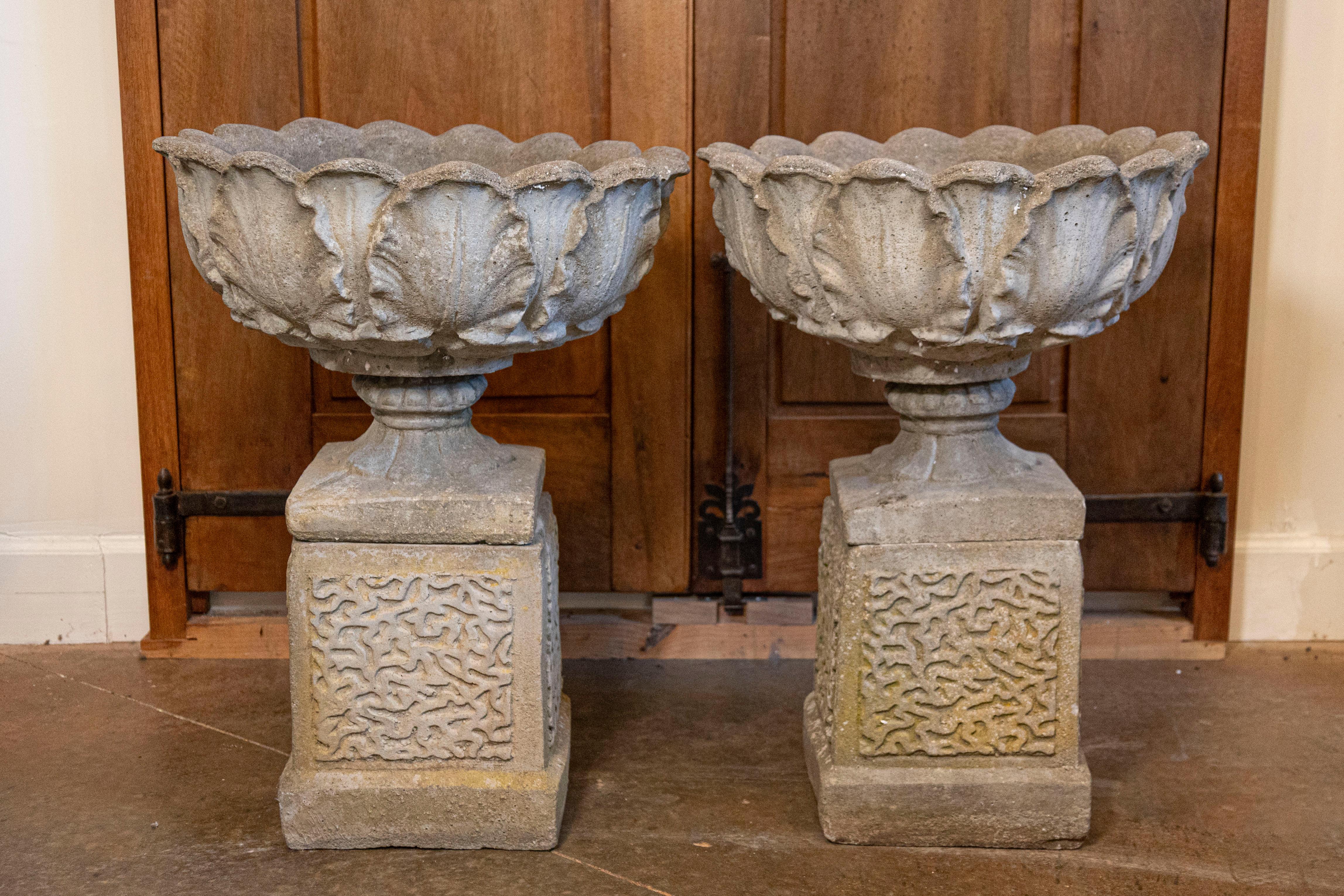A pair of English carved stone urns on pedestals from the 20th century, with acanthus leaf motifs. Created in England during the 20th century, each of this pair of stone urns captures our attention with its elegant rhythm of acanthus leaves adorning