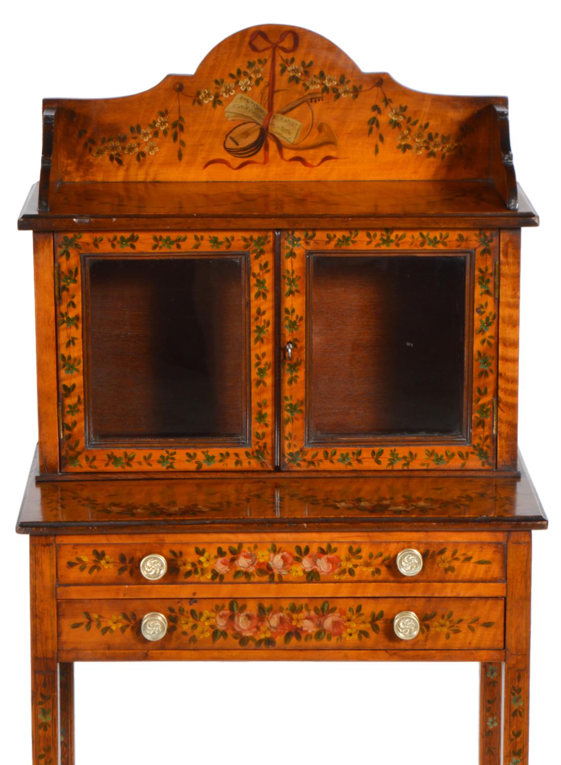 These 19th century slender display cabinets en chiffonier feature honey colored satinwood painted with flowers, ribbons and classical themes on all three sides. The long tapering legs are joined by a lower shaped shelf and supporting the upper part