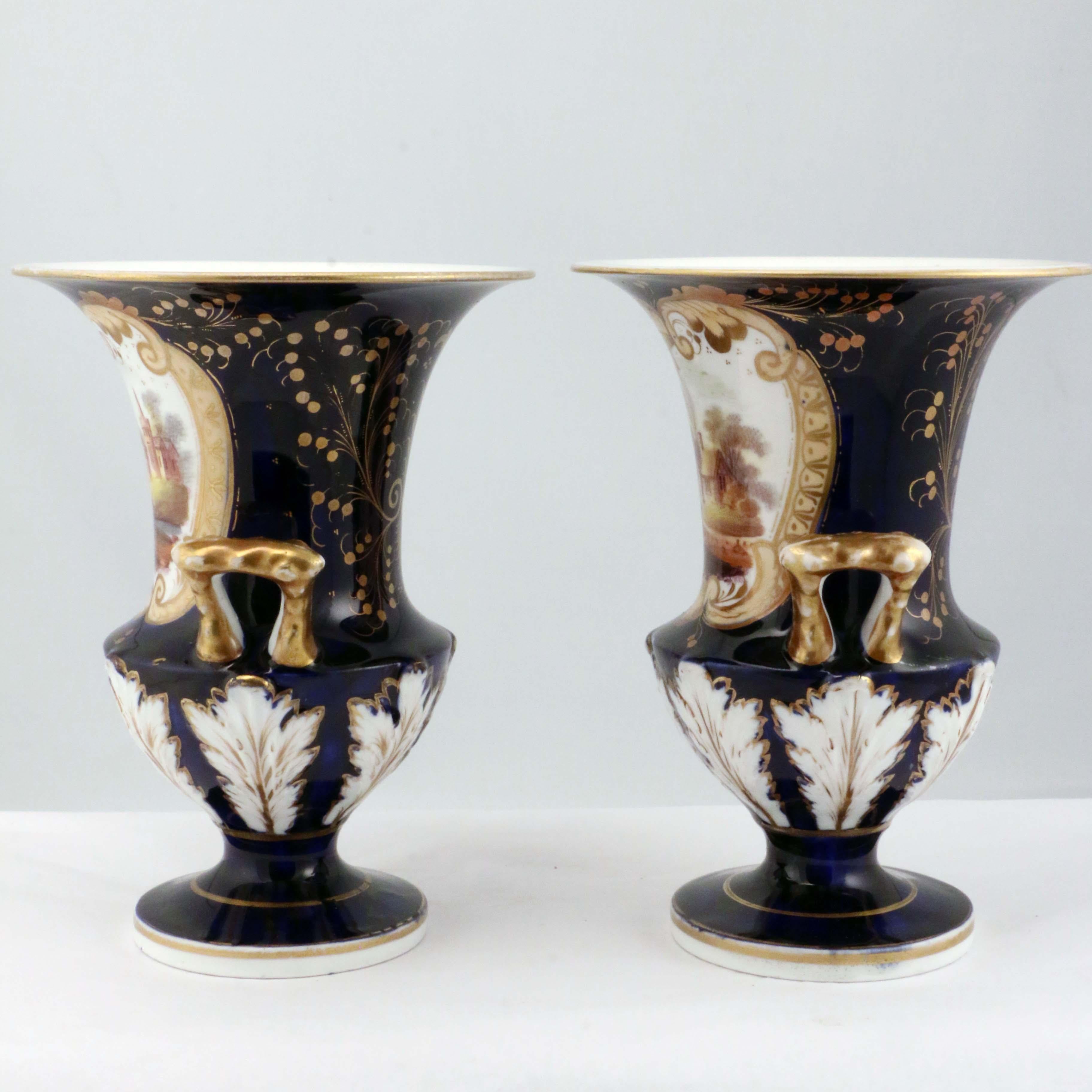 Each two handled urn is painted with a romantic view, one a castle, the other a country cottage, within a shaped gilt and yellow border. on a molded and cobalt-blue ground. The back is gilt with a stylized design, the base is molded with