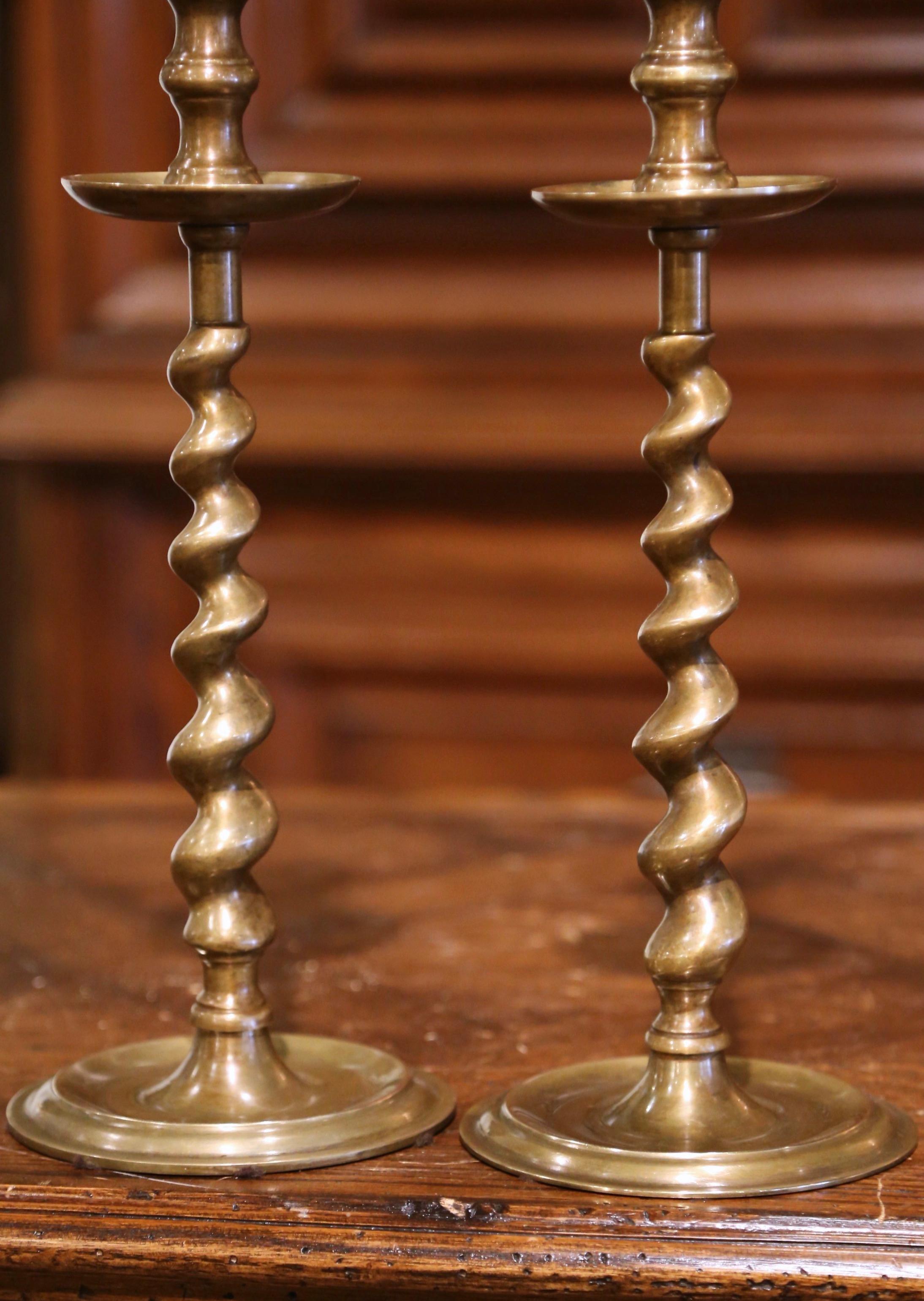 Decorate a dining table or console with this elegant pair of candlesticks. Crafted in England circa 1980, Each candle holder stands on a round vase over a barley twist stem. The lights are in excellent condition and adorn a rich patinated brass
