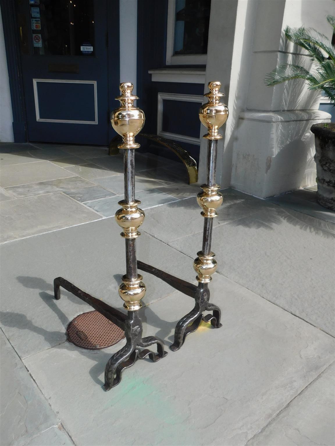 Pair of English brass and polished steel urn finial fire place andirons with decorative hand chase work, original wrought iron rear dog legs, and resting on scrolled legs with stylized penny feet, Late 18th century.