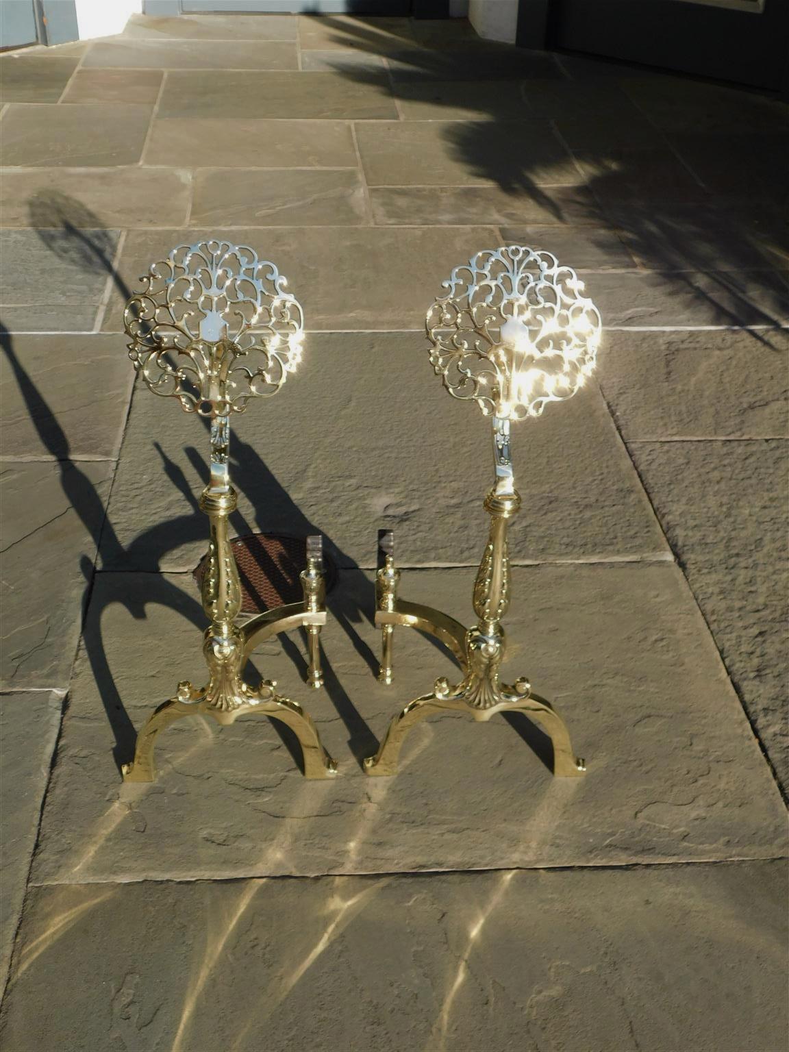 Pair of English cast brass flanking foliage pierced medallion andirons with centered acanthus plinths, matching urn finial log stops, and resting on scrolled legs with stylized flared feet. Stamped England on reverso. Mid 19th century.