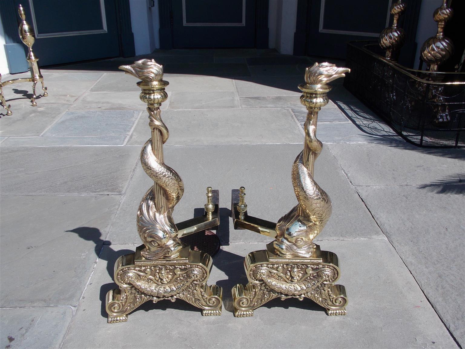 Pair of English brass intertwined dolphin andirons with flanking flame finials, matching urn finial log stops, original cast iron dog legs, and terminating on scrolled gadrooned legs with swag and acanthus motif. Early 19th century.
