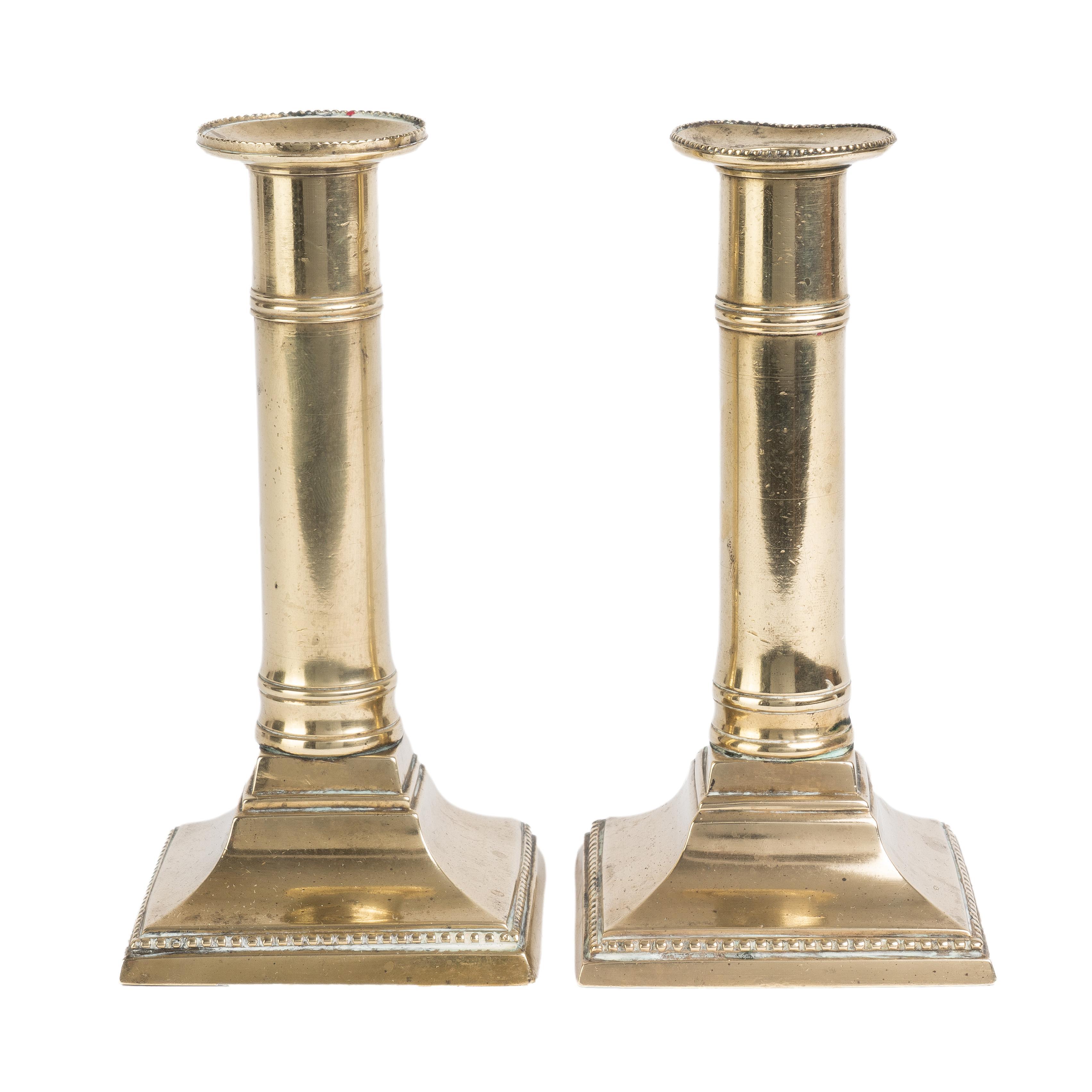 Pair of cast brass columnar candlesticks on a square base fitted with an internal push up candle ejector. Both the bobesh rim and the base are decorated with a finely beaded border.

England, circa 1810.