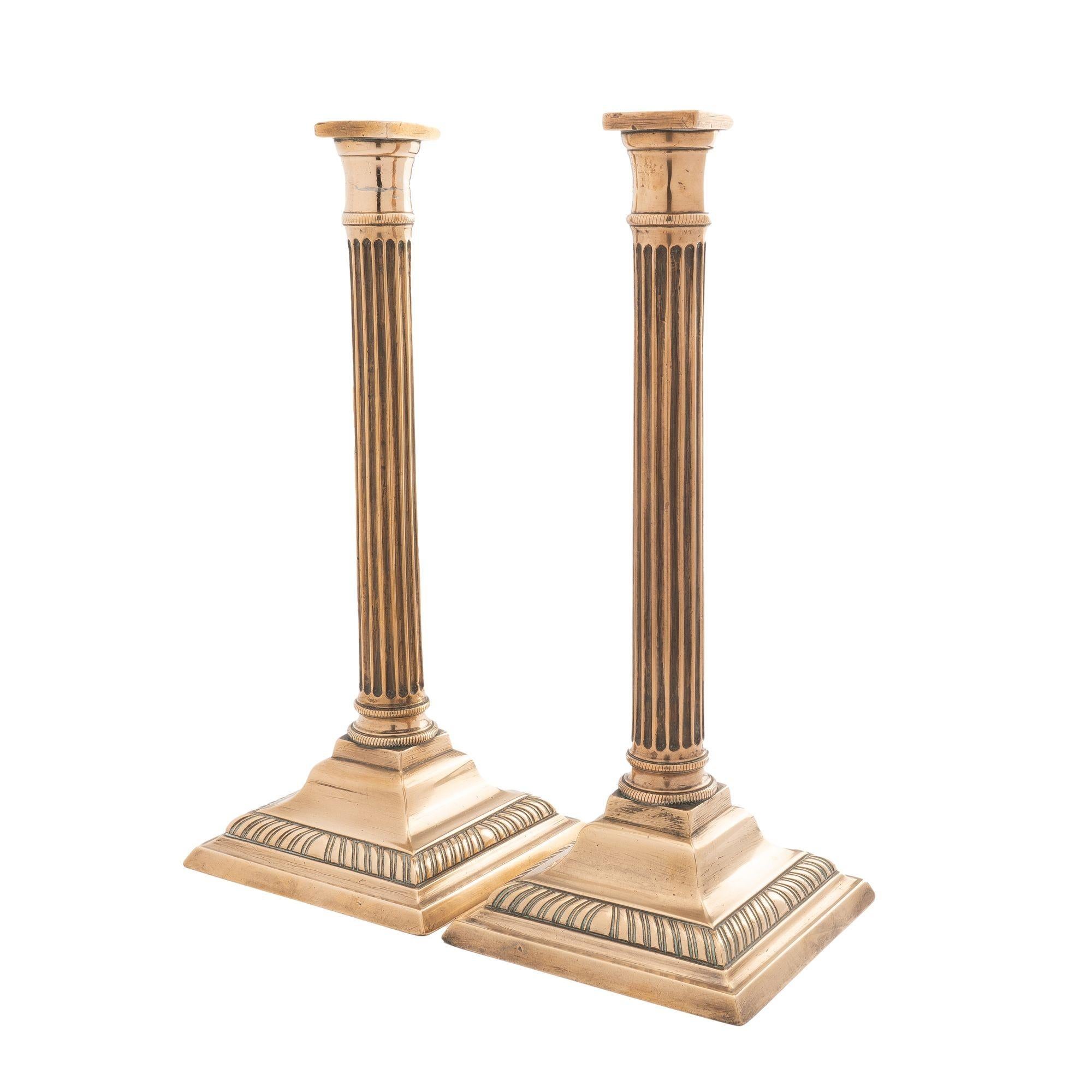 Pair of fluted columnar bell metal candlesticks on square stepped base with a gadroon molding. The hollow core cast candle shaft is peened to the square cast brass base. The fluted shaft incorporates a slightly concave circular candle cup with a