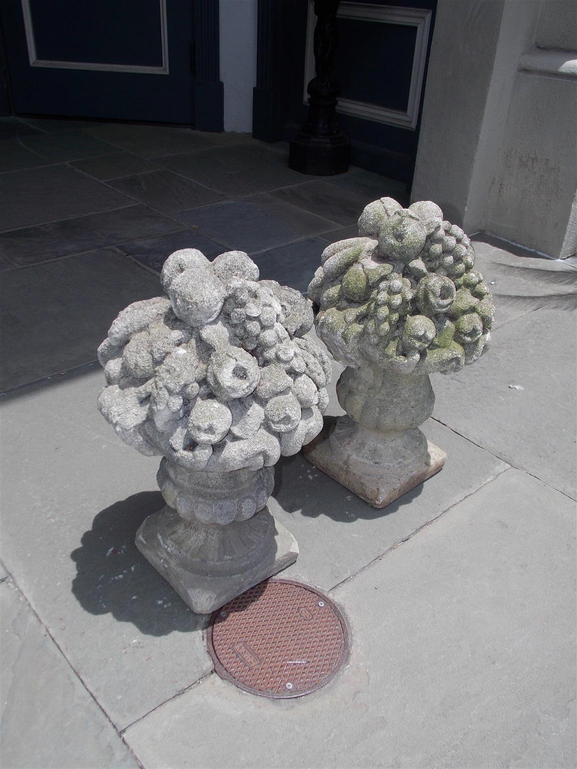 Pair of English cast stone floral and fruit basket garden ornaments resting in flanking gadrooned camping urns, and terminating on circular fluted squared plinths, Early 19th century.