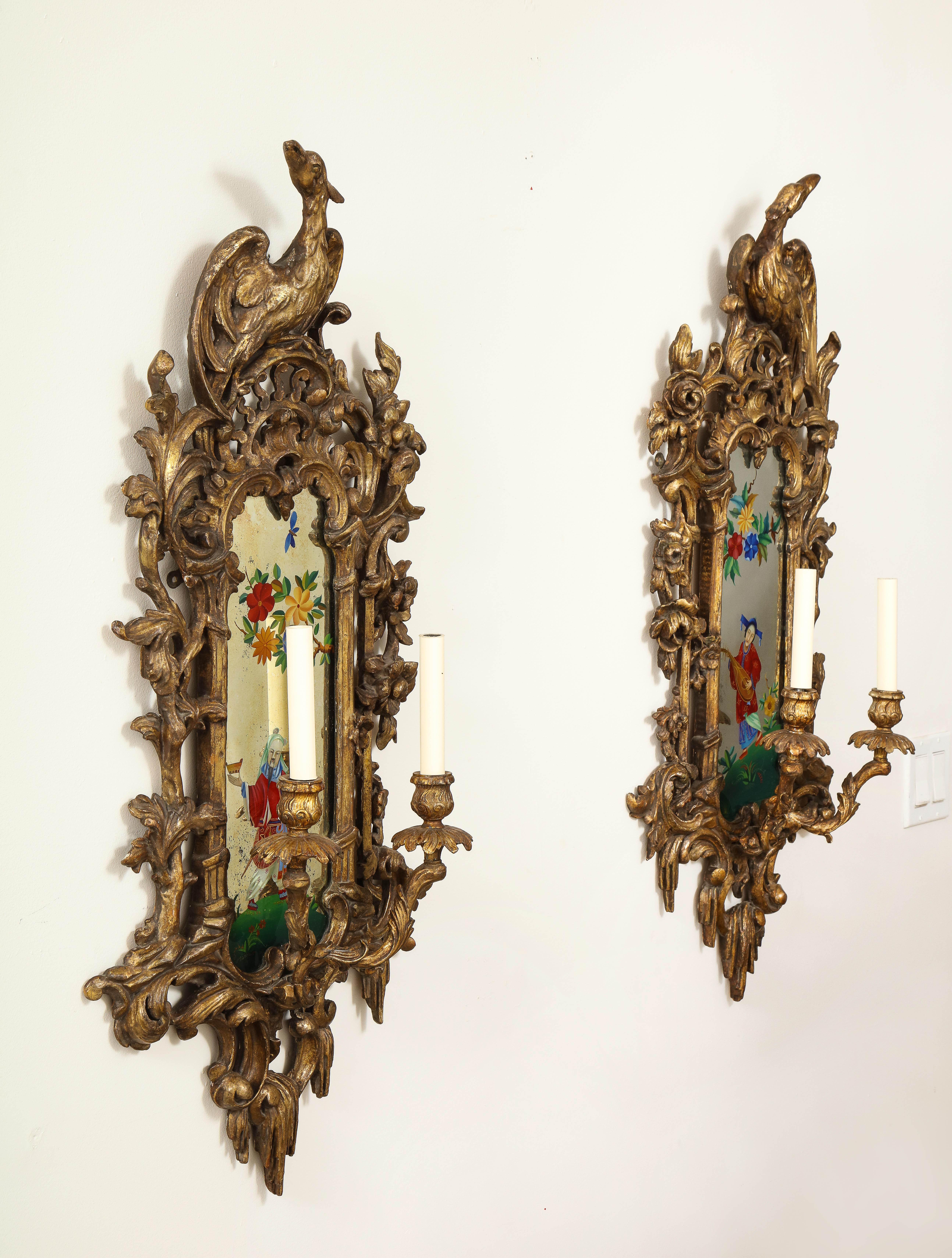 A fantastic pair of English chinoiserie painted reverse on glass, carved giltwood mirrored two-light sconces. Each is beautifully hand carved and gilt in a gorgeous gold finish with fabulous hand carved open-work and two mirror-image giltwood birds