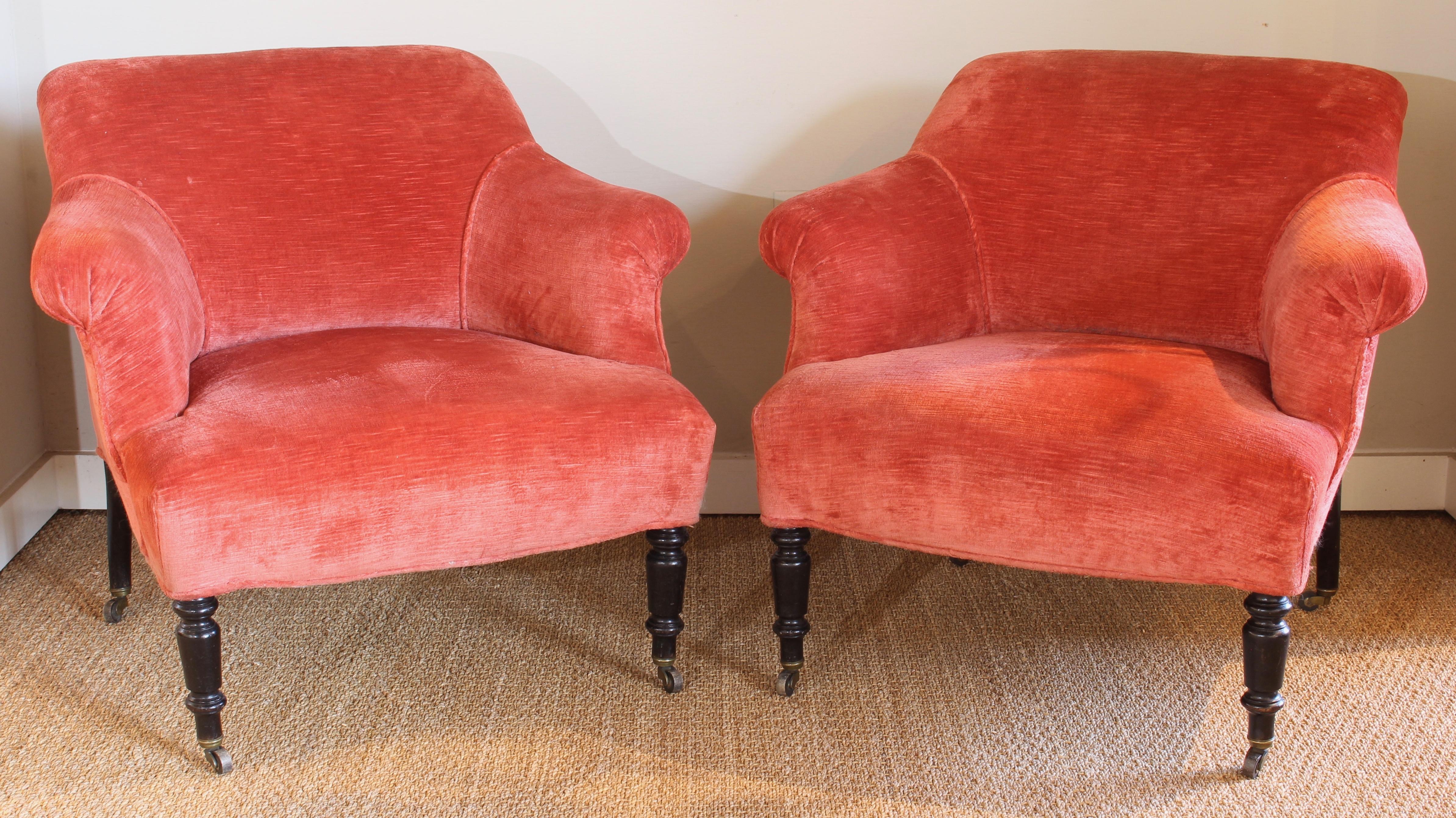 A pair of late 19th century small English club chairs covered in a deep coral linen velvet resting on turned ebonized legs terminating in brass casters.
