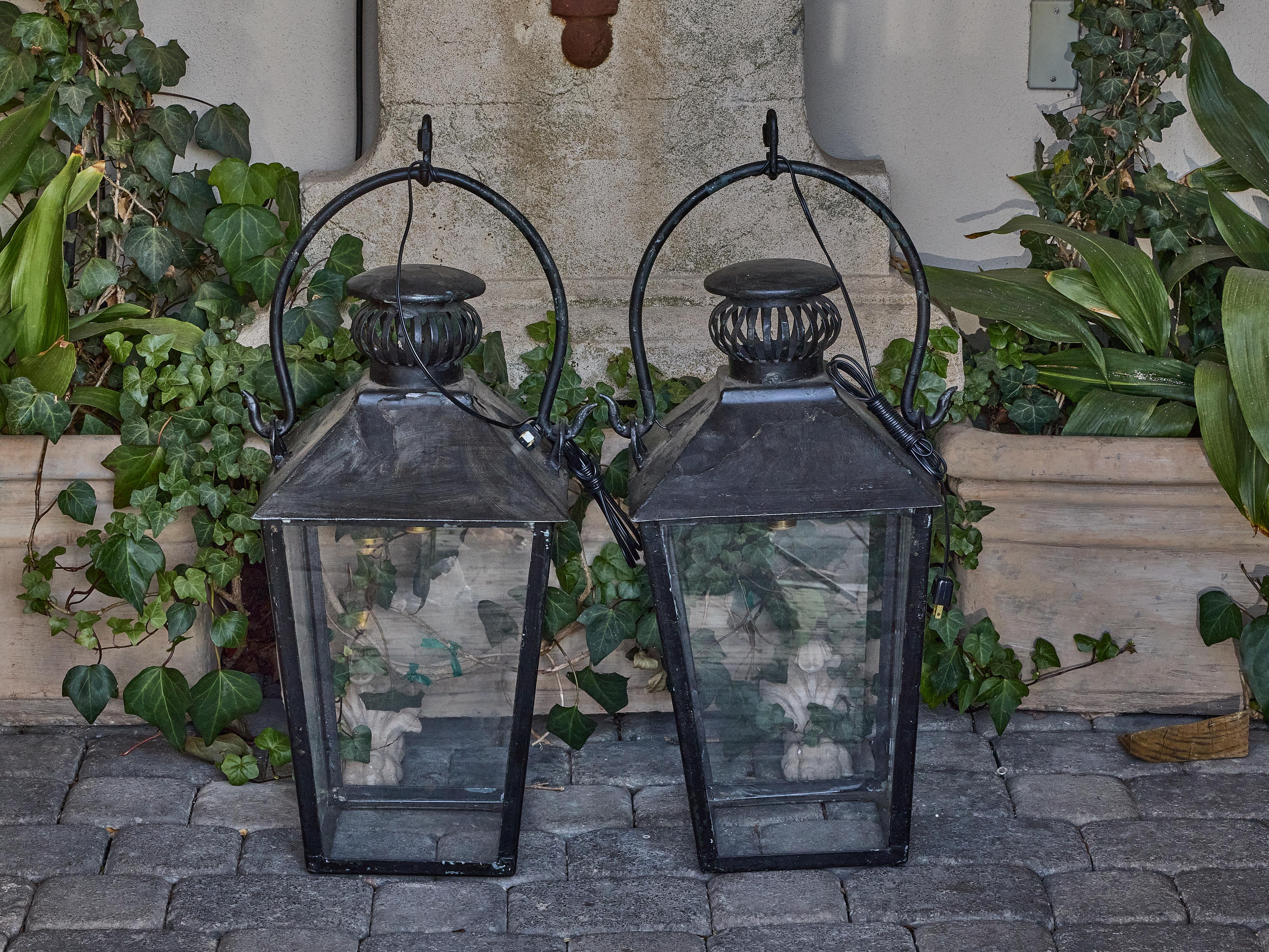 Cette paire de lanternes en fer vintage apporte un charme anglais classique à tout espace extérieur ou intérieur couvert. Fabriquée au milieu du XXe siècle, chaque lanterne présente une silhouette effilée avec un cadre à quatre côtés et des panneaux