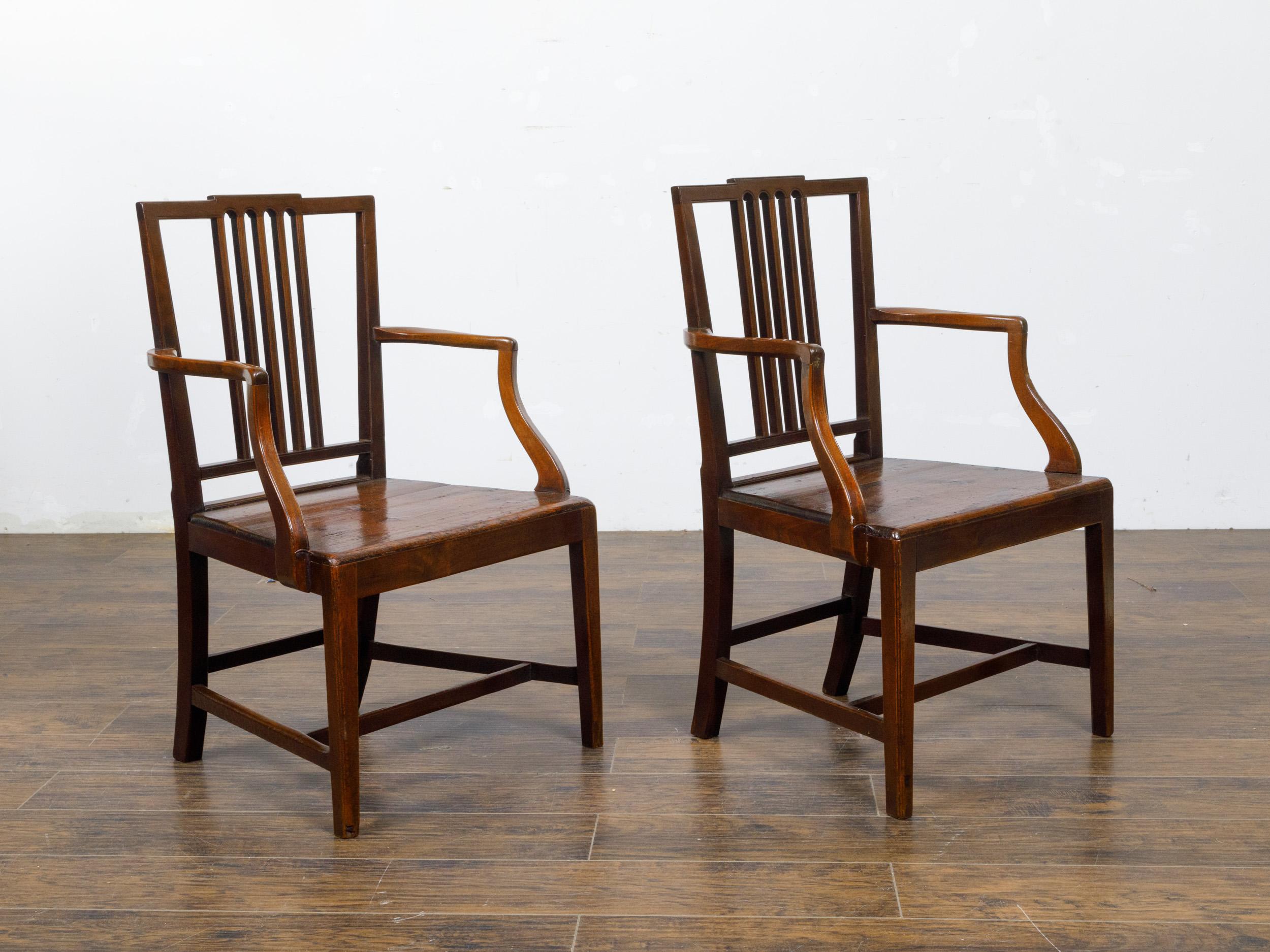 A pair of English George III period plank seat chairs from circa 1800 with pierced backs, slightly scrolling arms, straight legs and cross stretchers. This pair of George III period chairs hails from early 19th-century England, showcasing the