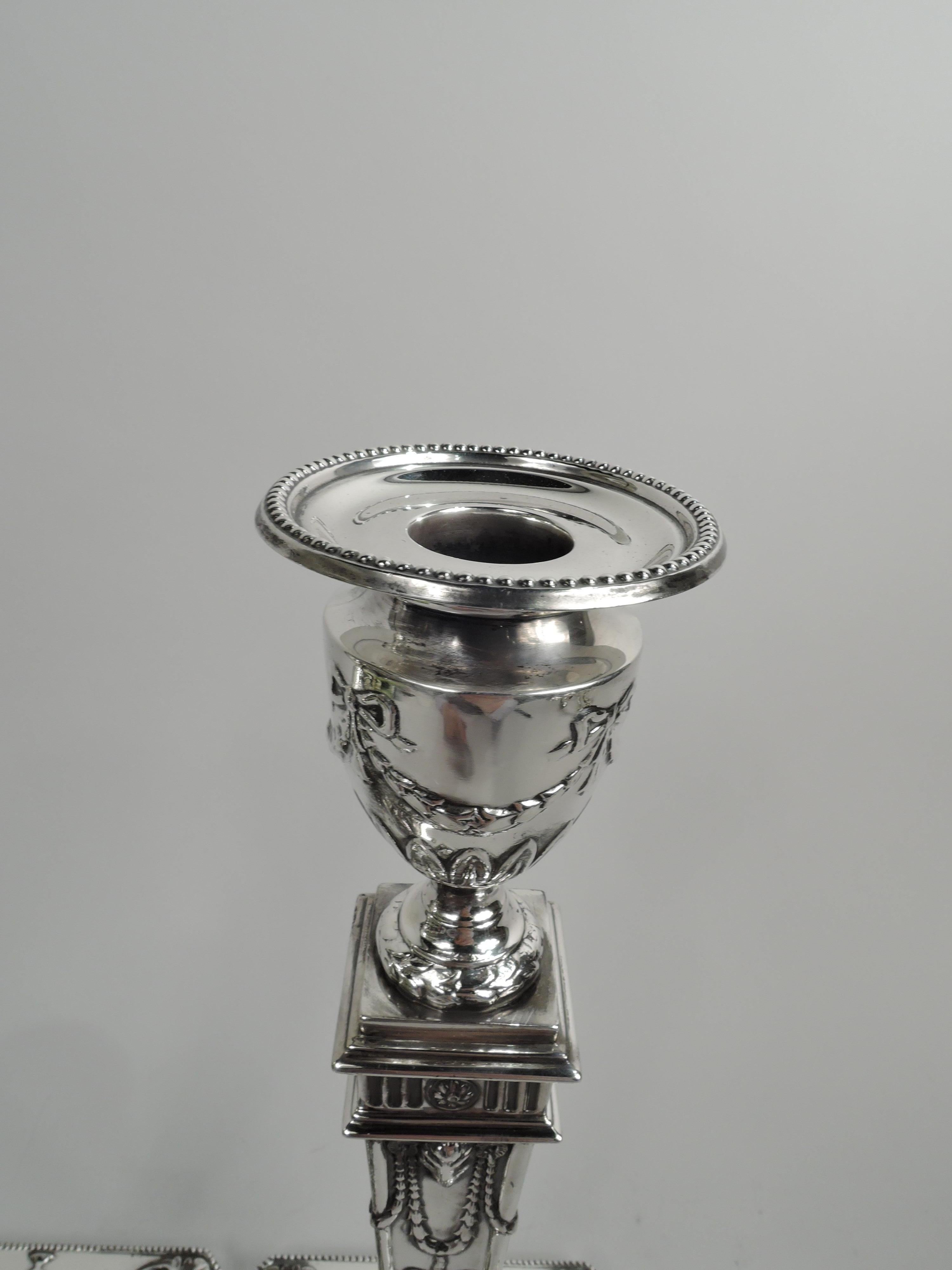 Pair of George V sterling silver candlesticks. Made by Ellis Jacob Greenberg in Birmingham in 1912. Each: Tapering pillar on raised and chamfered square base. Urn socket with detachable bobeche. Ram’s heads, ribboned garlands, paterae,