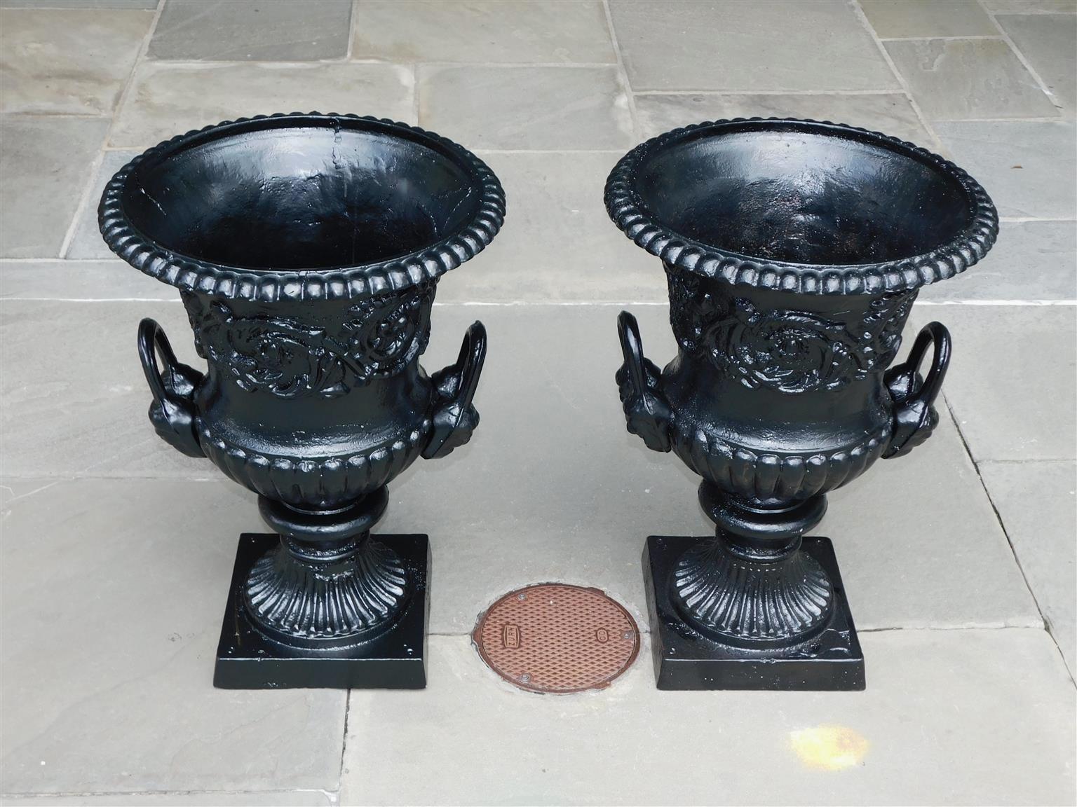 Pair of English figural cast iron and painted campana-form urns with an intertwined floral and medallion frieze, flanking figural double side handles, gadrooned motif, and terminating on circular fluted squared plinths, late 19th century. Pair have