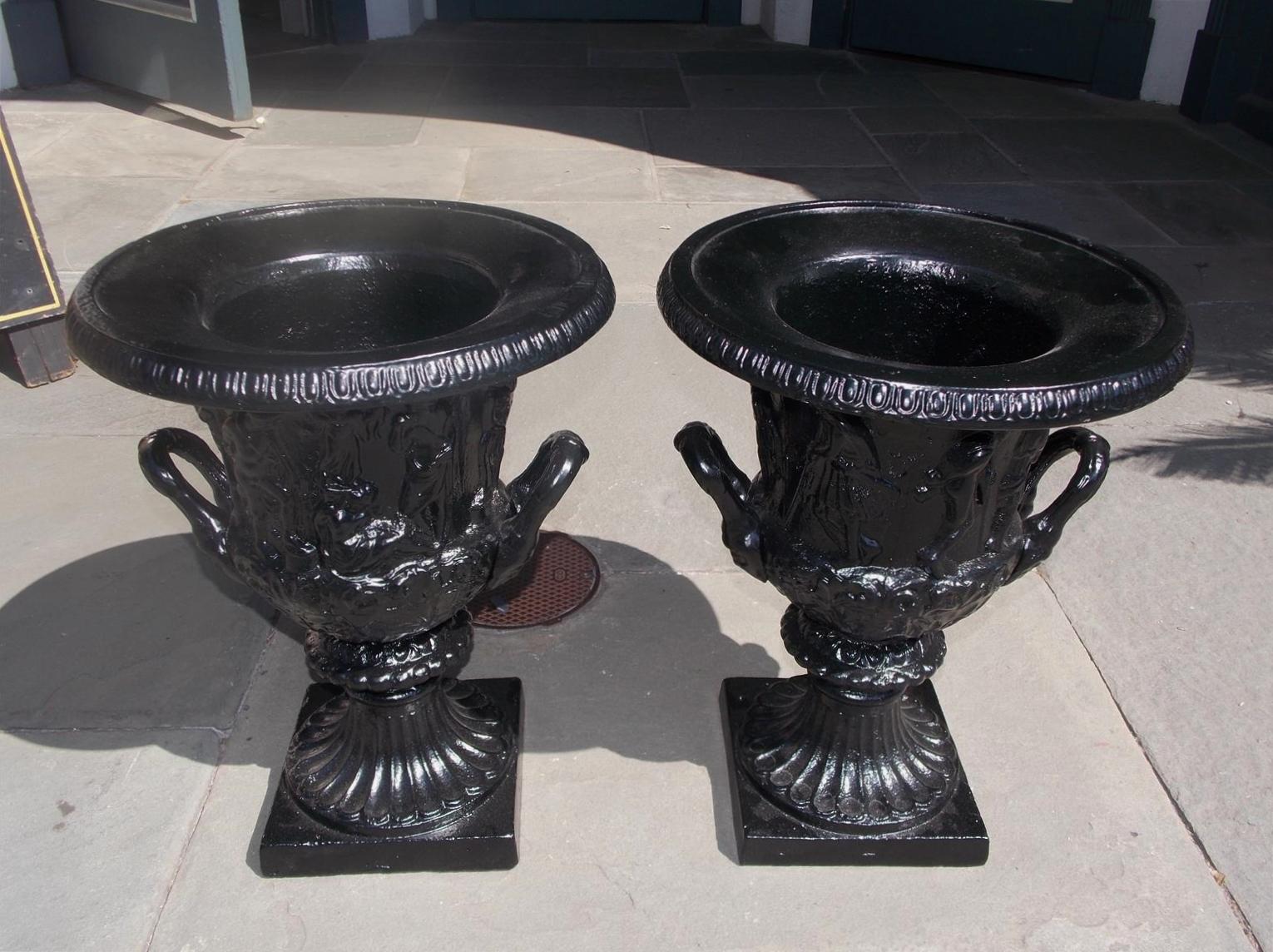 Pair of English figural frieze cast iron powder coated Charleston green campana-form urns with flanking side handles, and terminating on circular fluted squared plinths.  Late 19th century.
Measures: 20 Tall / 15 Diameter top / 8.5 squared base. 
 