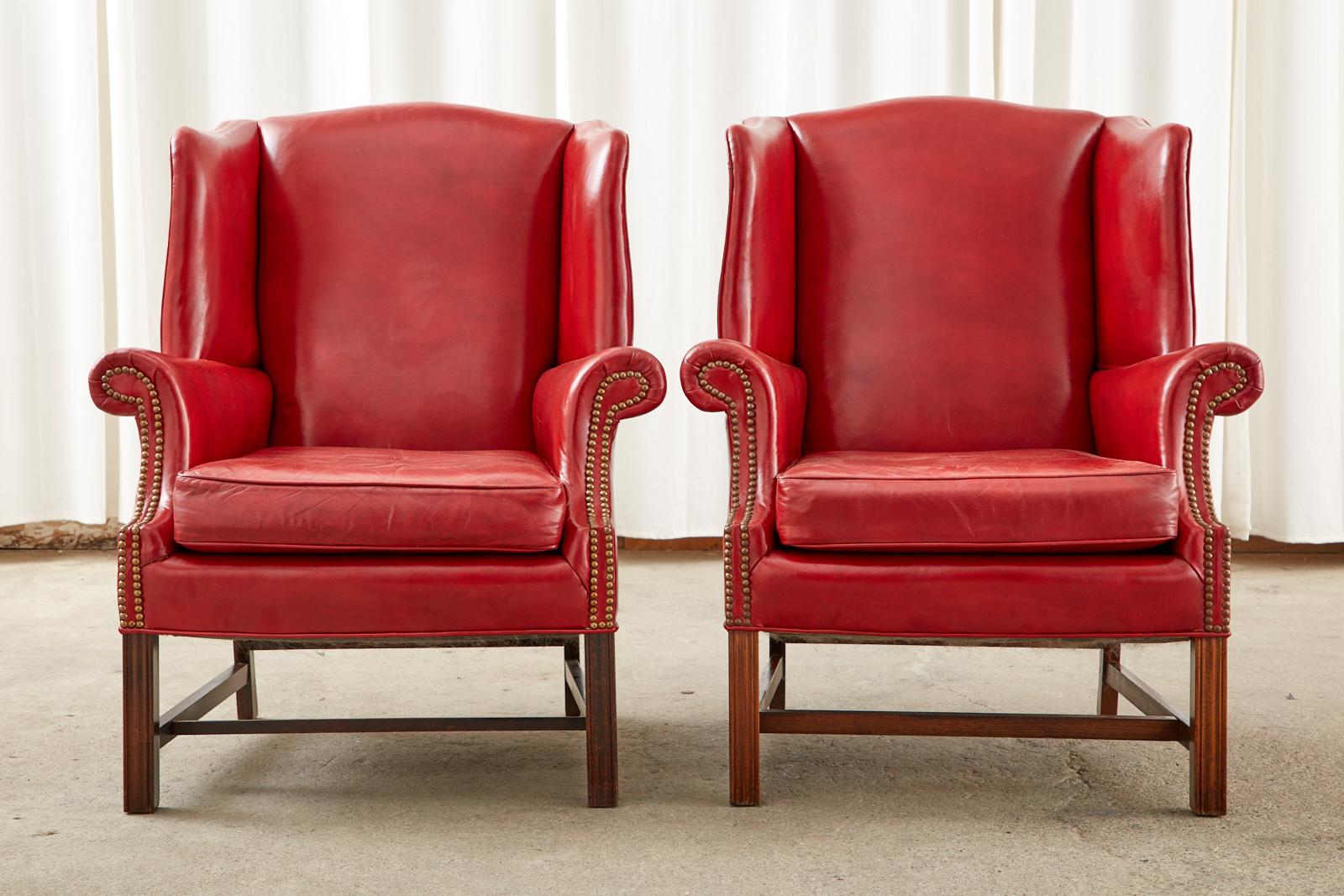 Stunning pair of mid-century leather wingback chairs or wing chairs featuring a dramatic ruby red leather upholstery. Made in the grand English Georgian taste with a mahogany hardwood frame. The chairs have large fully developed wings conjoined to