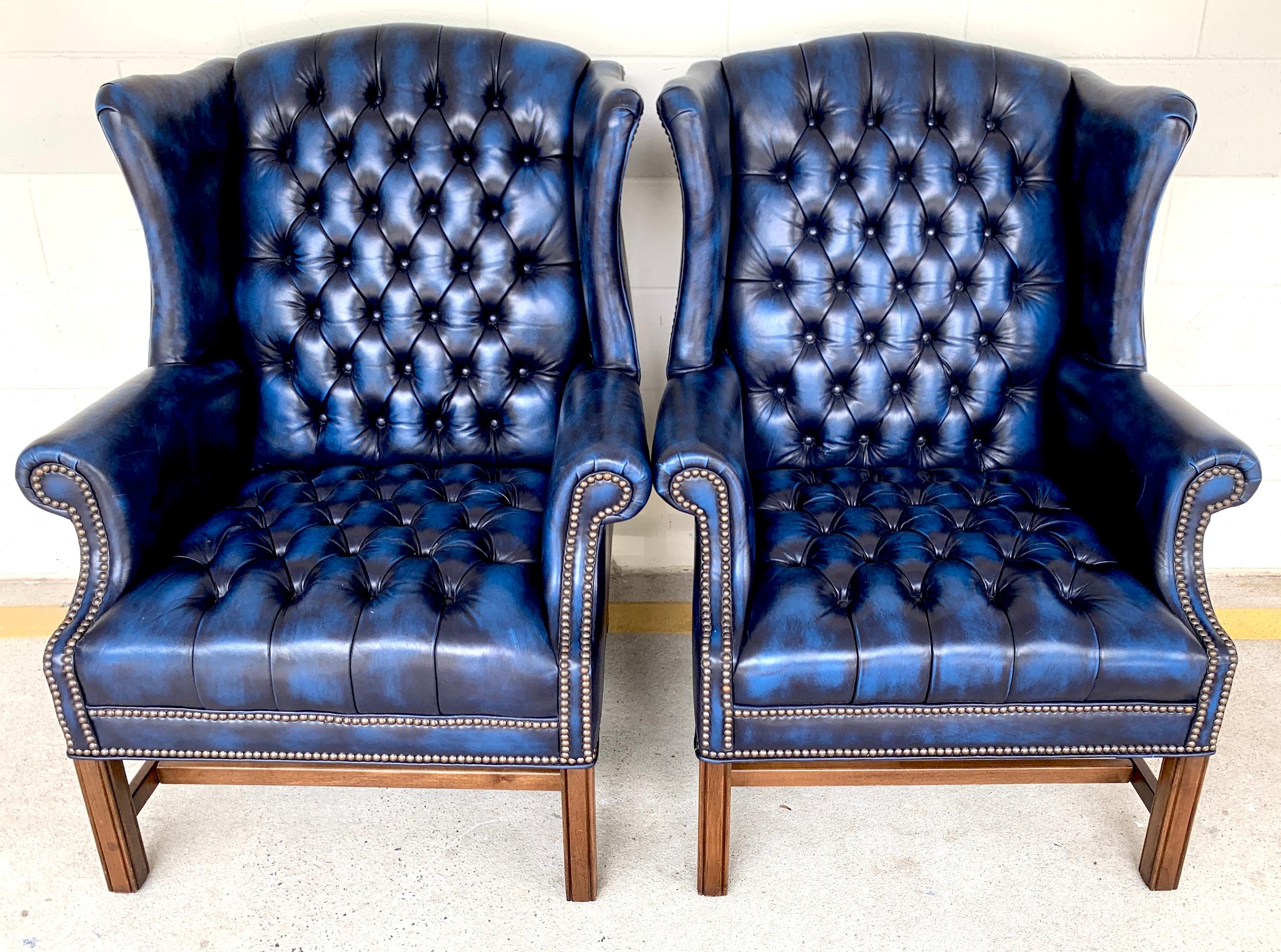Pair of English Hollywood Regency blue leather wing back Chesterfield chairs, each one with tufted and nailhead detail, custom hand dyed shaded blue leather upholstery raised on four mahogany Marlborough legs, with 'H' stretcher. Clean and simple